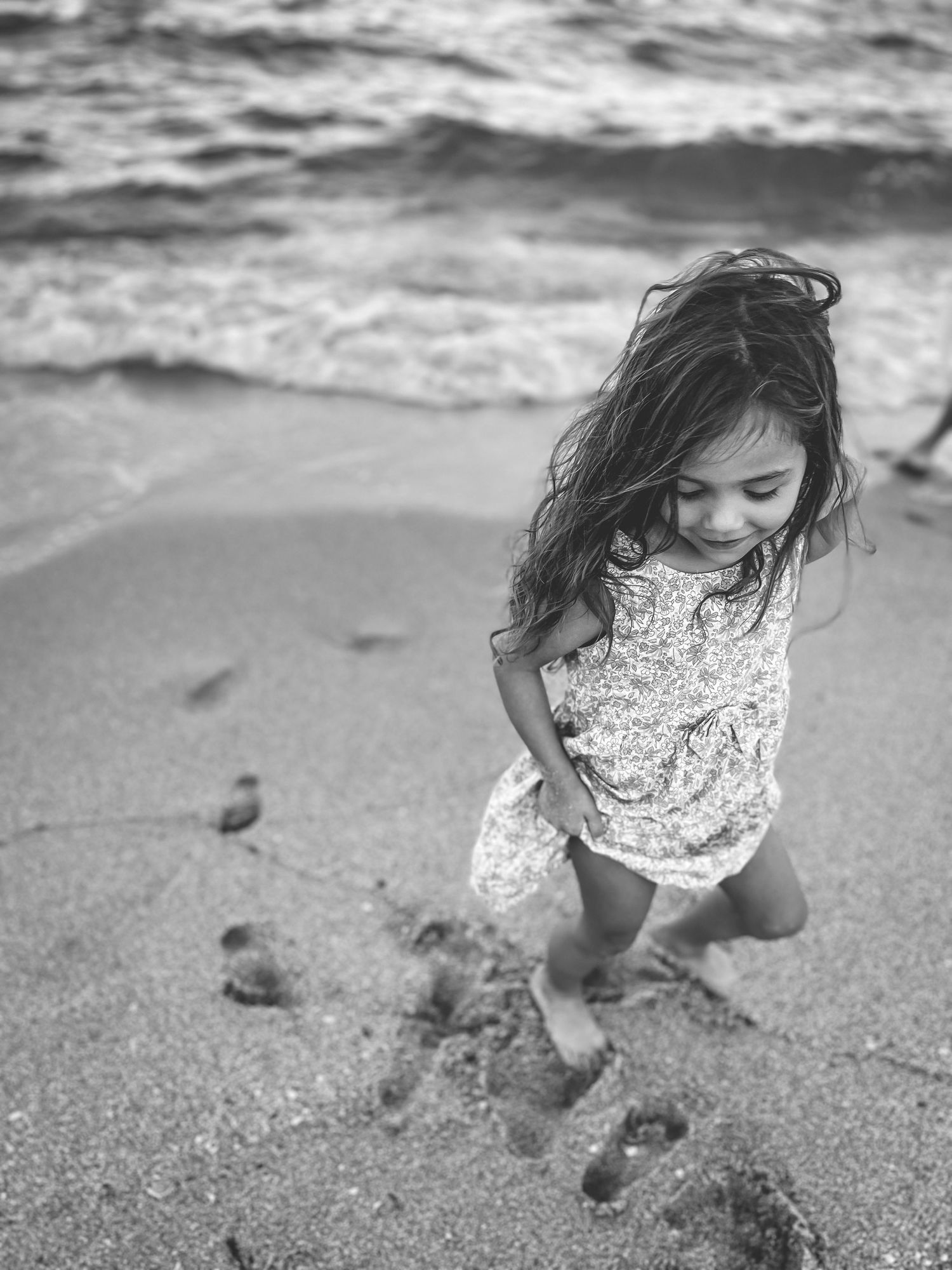 A playful child in a floral dress runs along the beach shoreline in black and white photography.