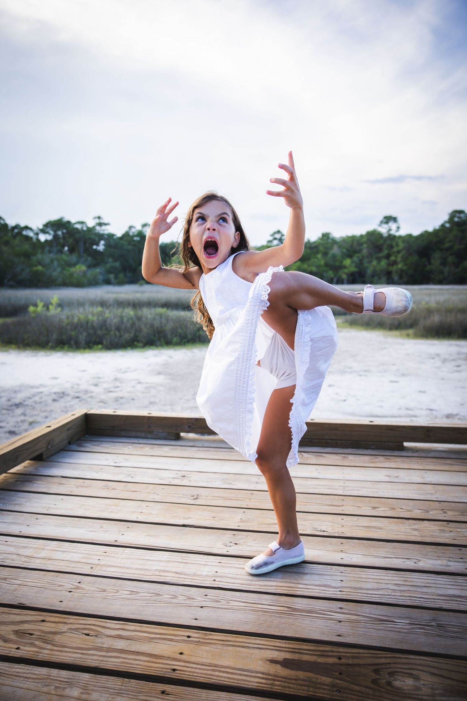 A joyful dancer in a white dress performs an energetic pose on a wooden dock by the water.