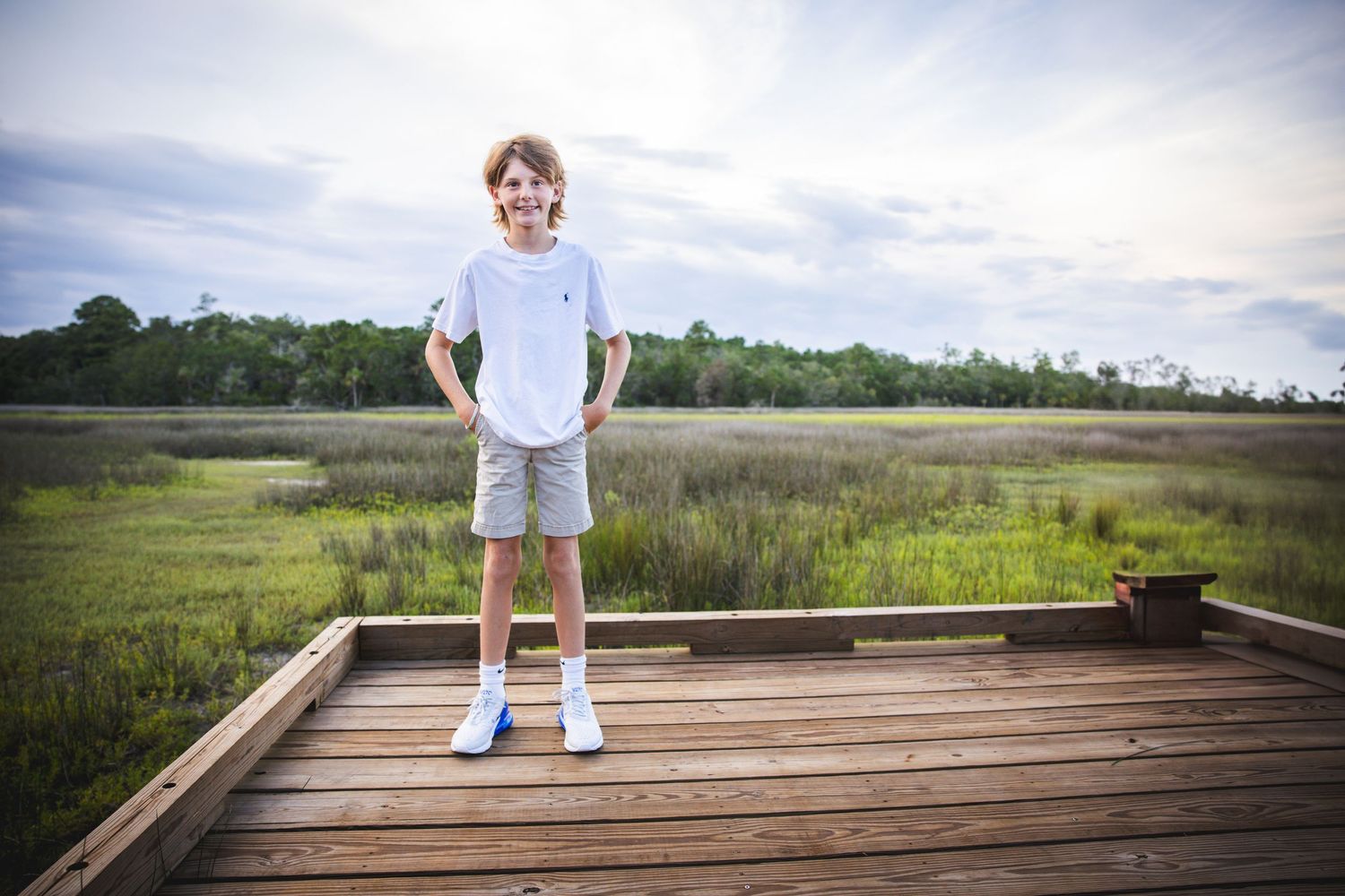 A wooden boardwalk extends through a serene marsh landscape under cloudy skies.