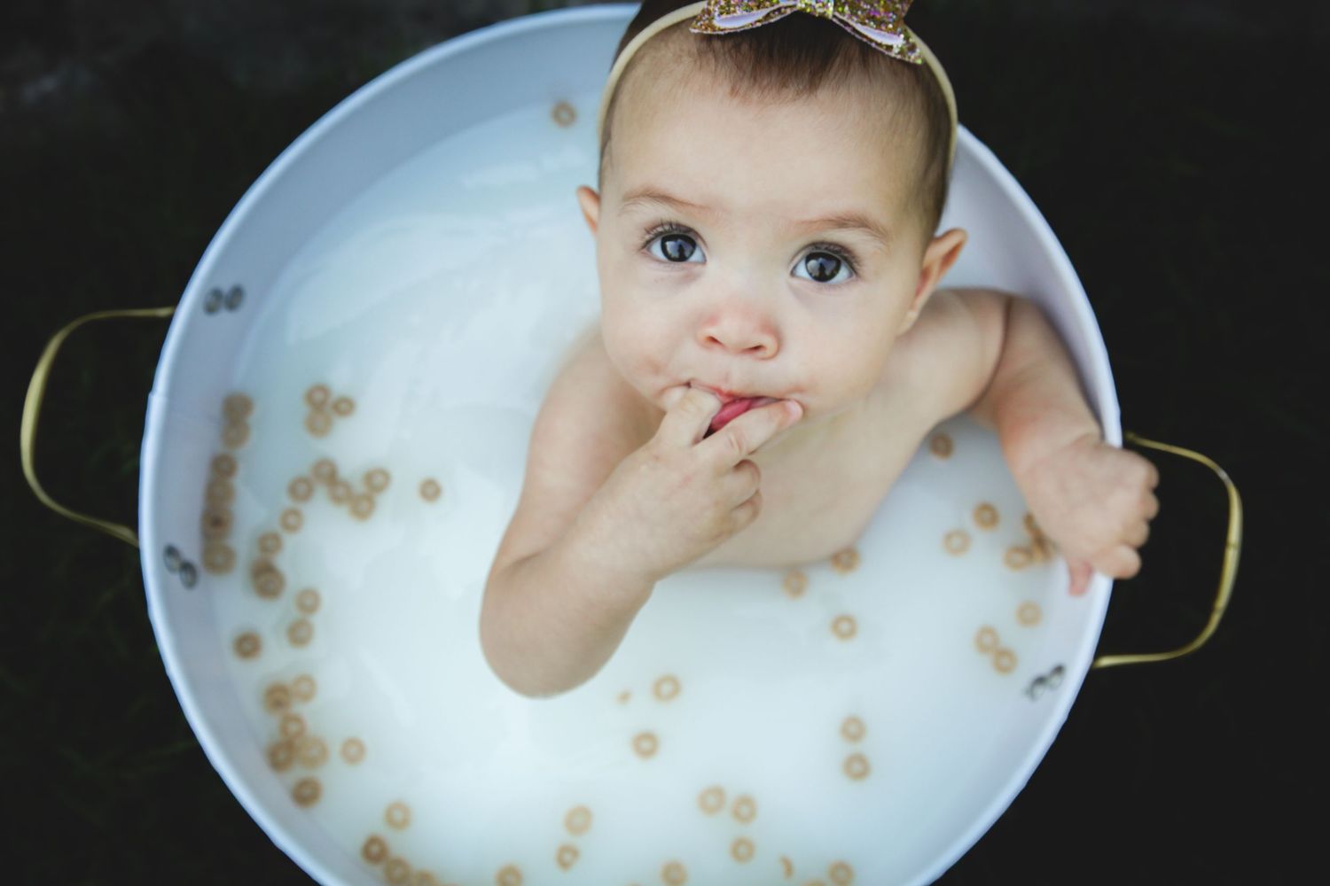 Baby sitting in white tub with Cheerios floating in milky water, photographed from above.
