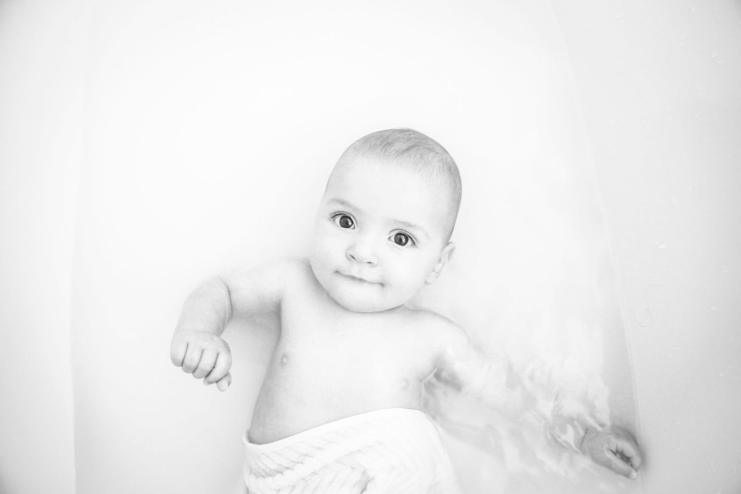 Black and white portrait of a smiling baby wrapped in a white cloth against a light background.