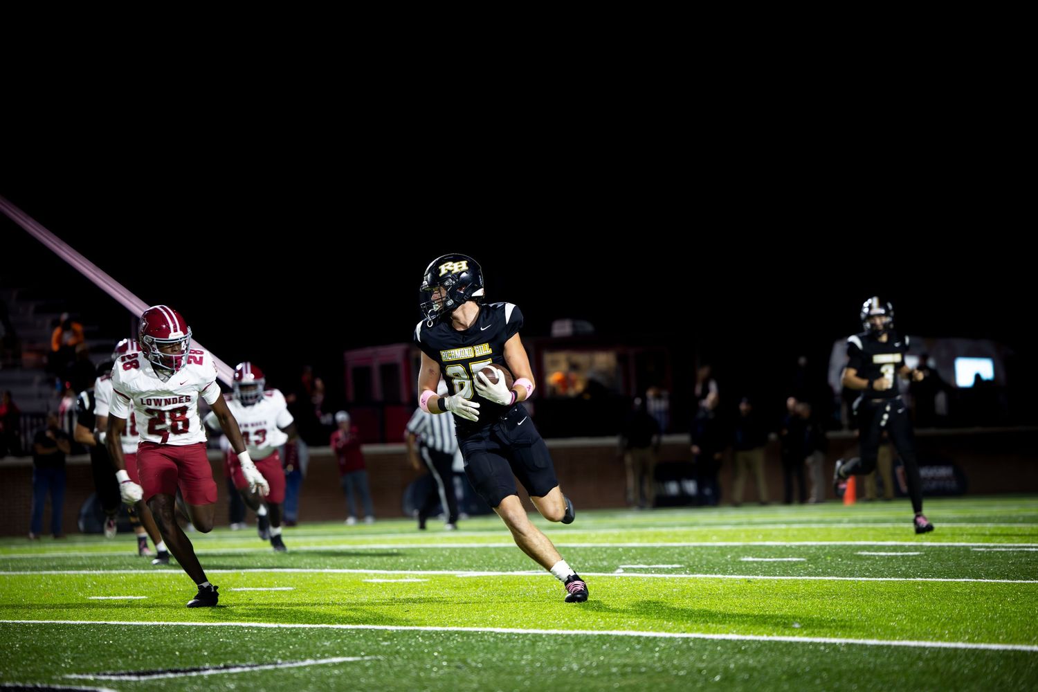 Night football game with players in dark uniforms running on field, pursued by opponent in red jersey number 26.