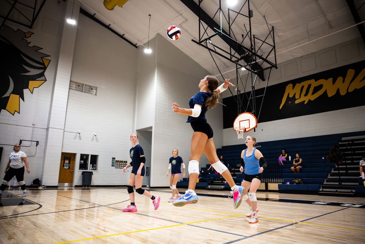 Female volleyball player jumping to spike the ball during indoor practice at Midway gymnasium with teammates watching.