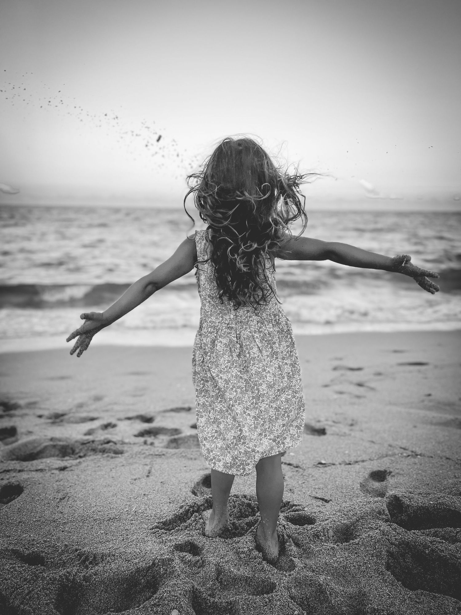A silhouetted figure in a floral dress stands with arms outstretched on a beach in black and white photography.