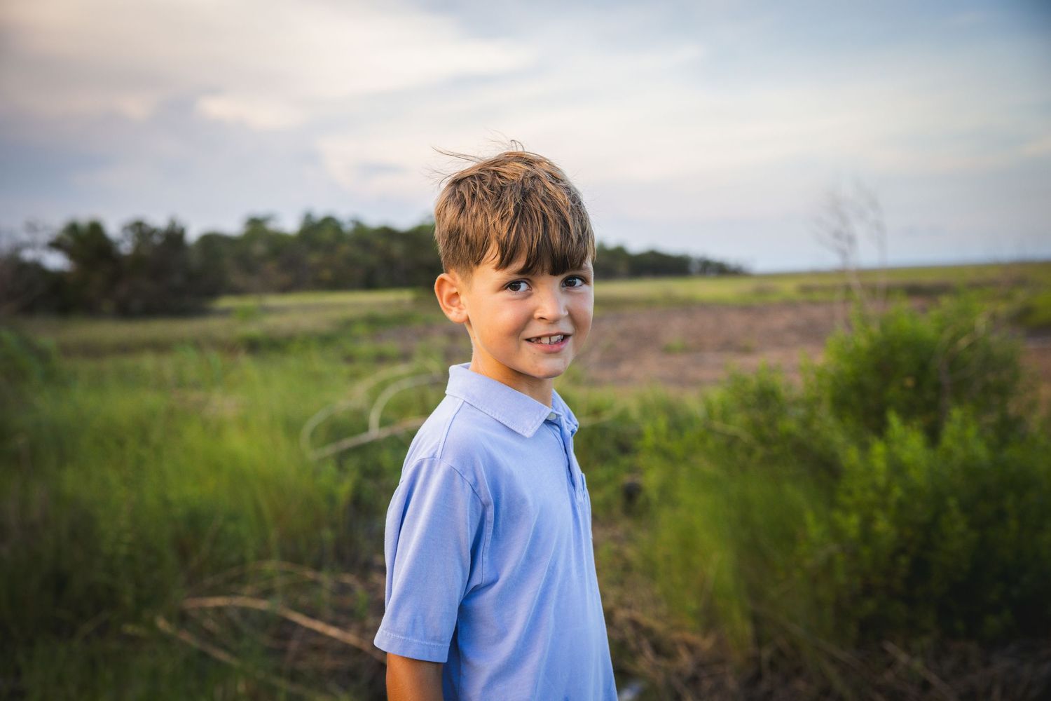 A smiling child in a blue shirt stands in a grassy field during golden hour.