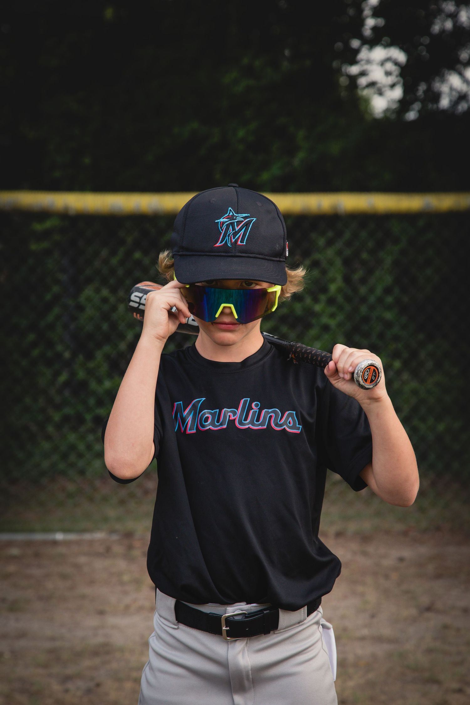 Young baseball player wearing a Miami Marlins cap and jersey adjusts sunglasses while standing on a baseball field.