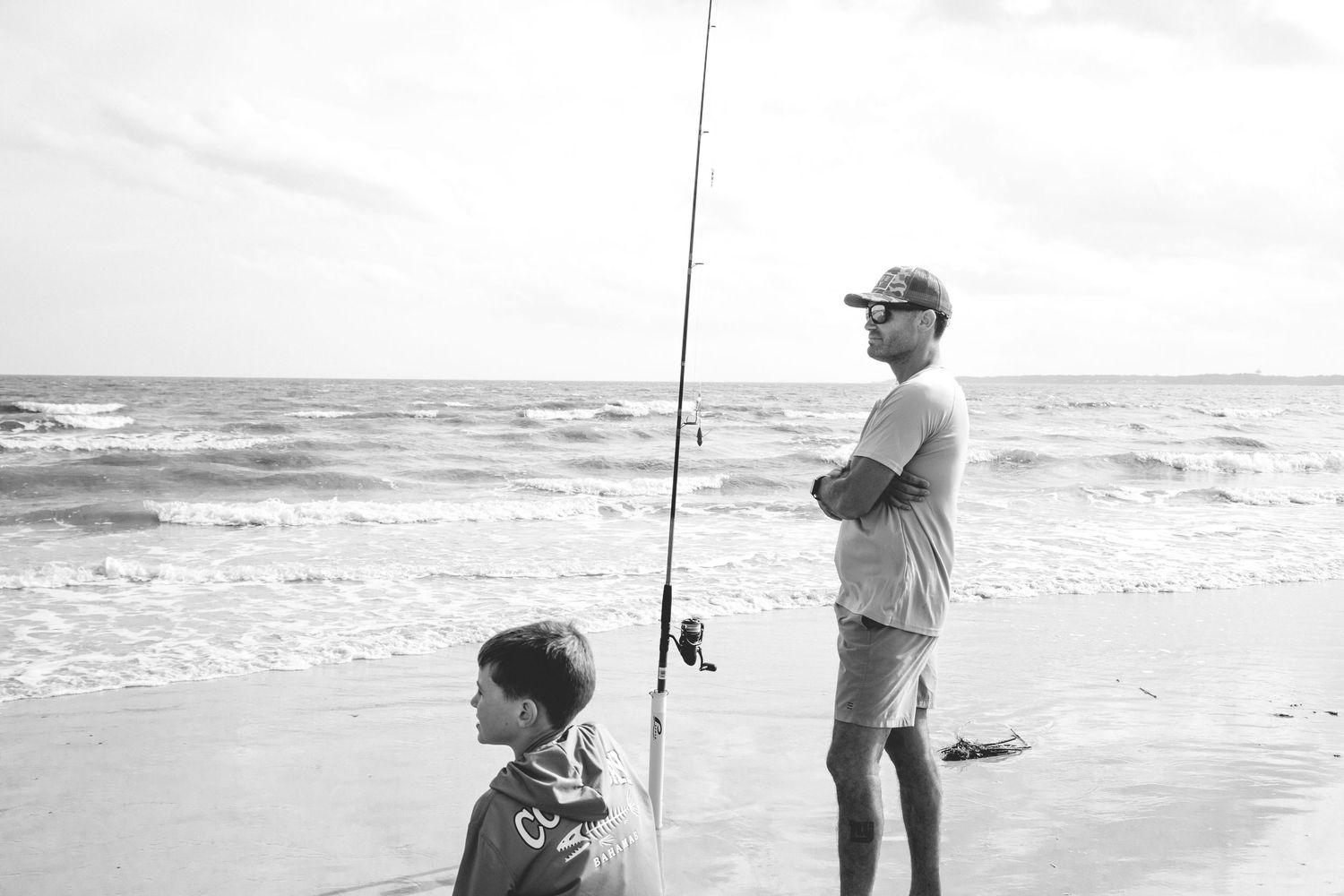 A black and white photo of fishing at the beach as waves crash onto the shore.