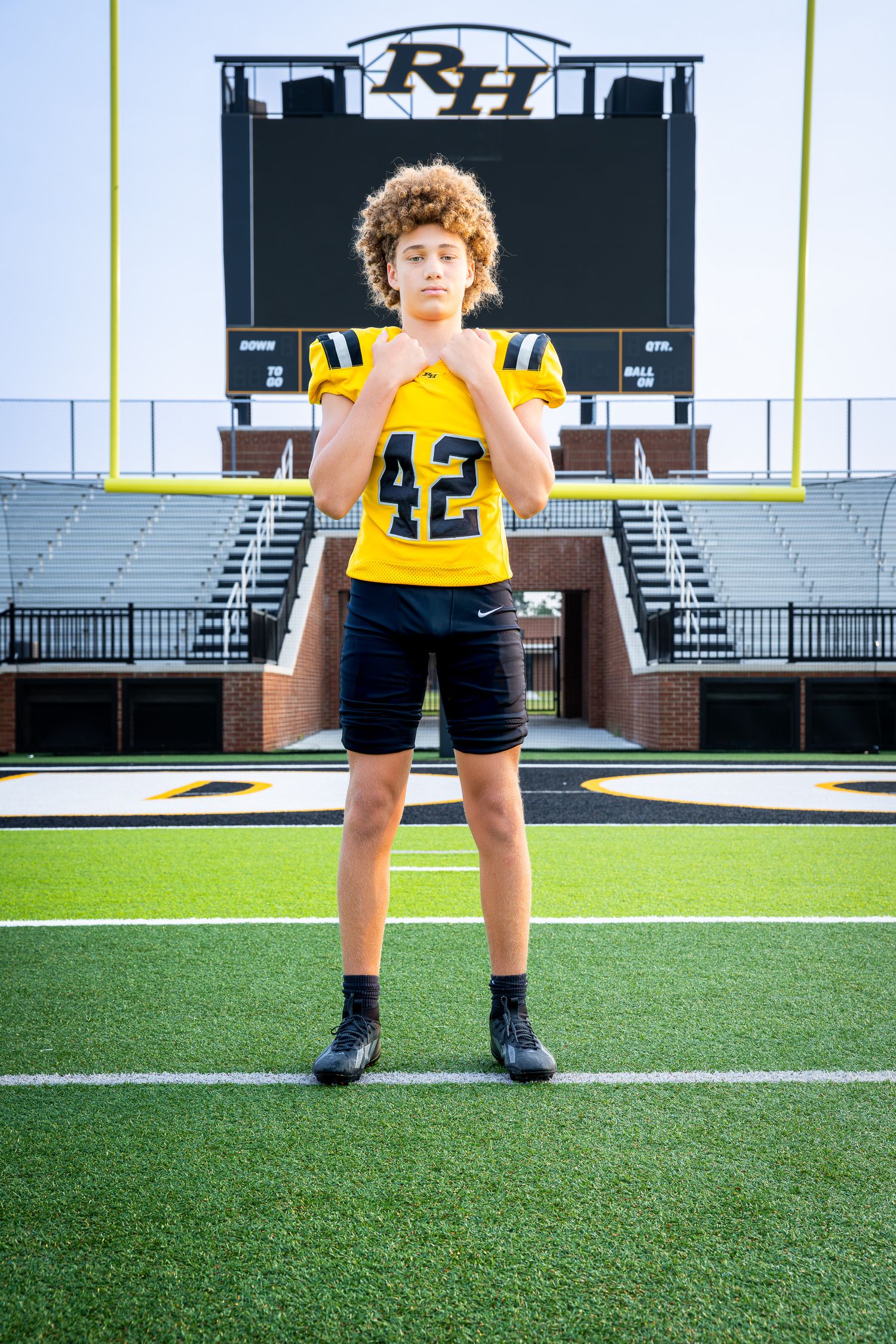 Young football player #42 in yellow jersey poses confidently on green turf field with stadium scoreboard behind him.