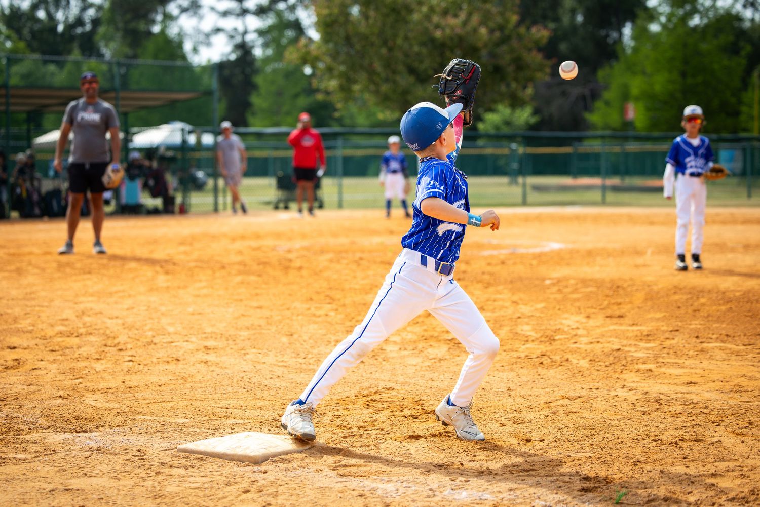 Young baseball player in blue uniform catching a ball at base during a little league game on a sunny day.
