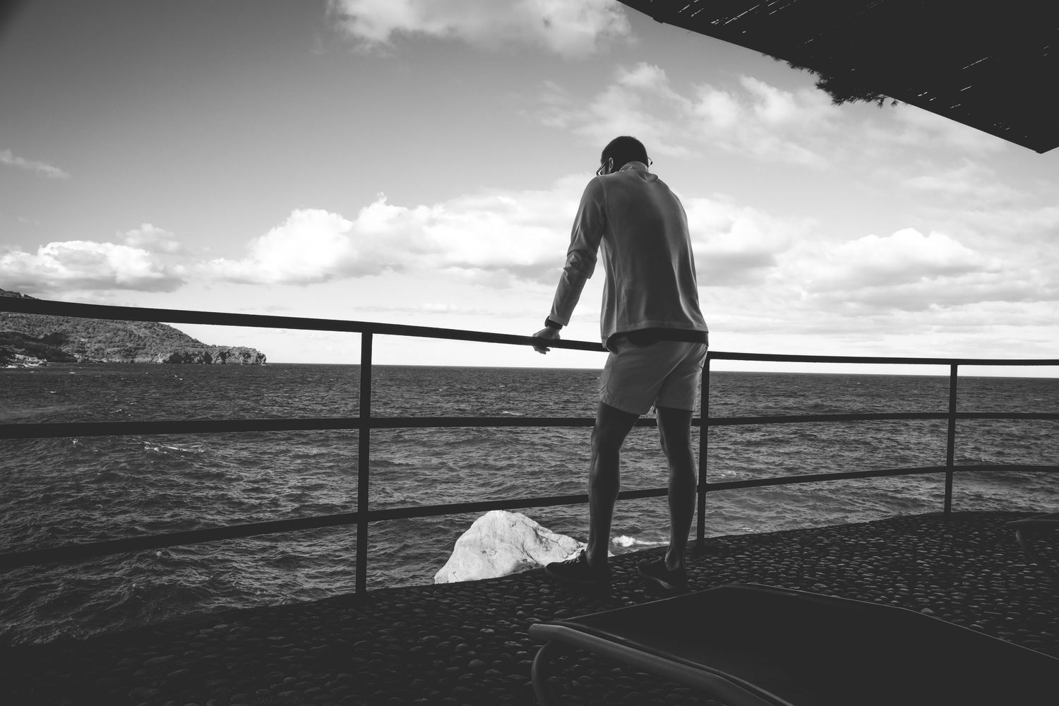 A series of black and white silhouetted figures stand along coastal railings against dramatic ocean views at sunset.