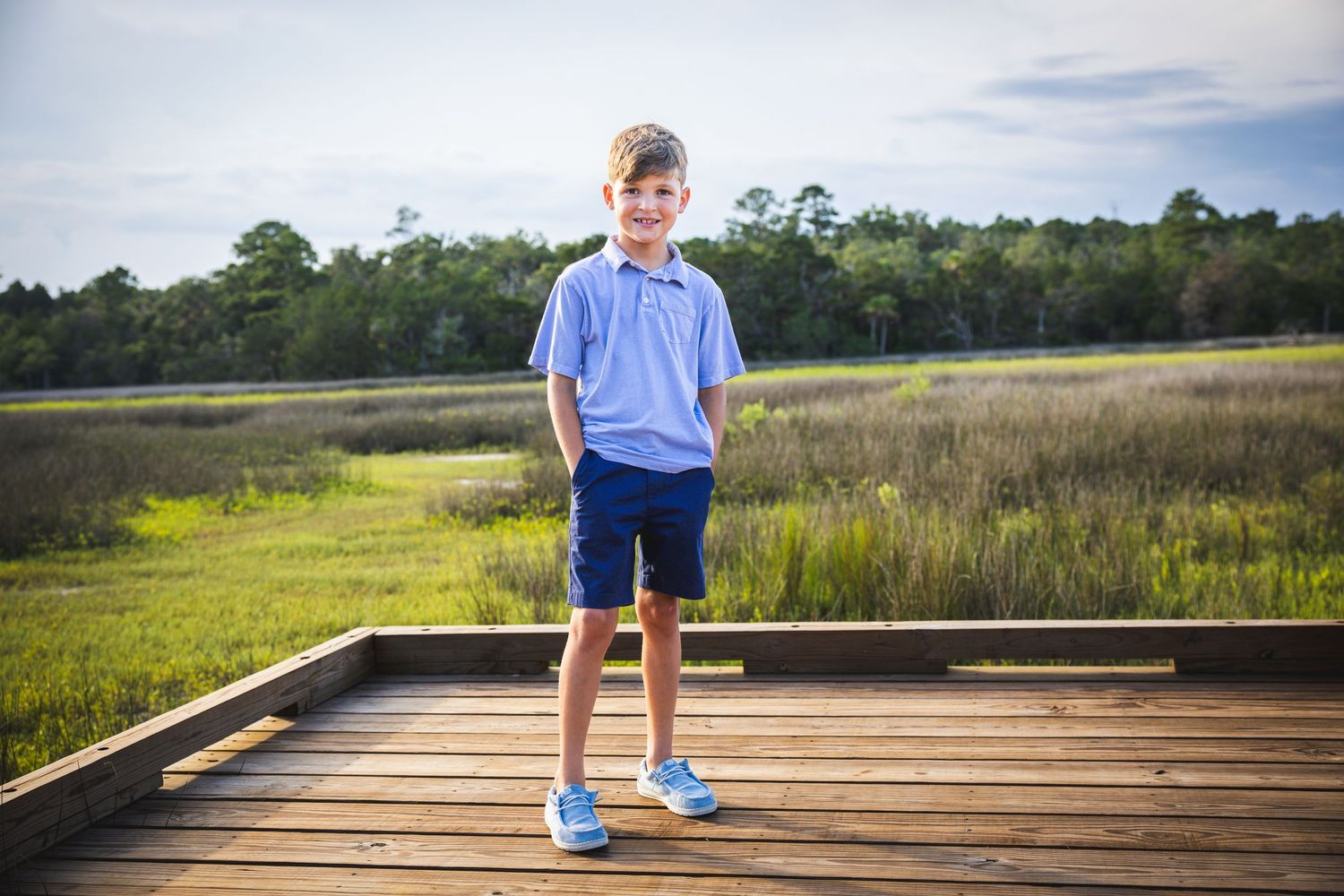Person in blue shirt and navy shorts standing on wooden deck overlooking marsh landscape.