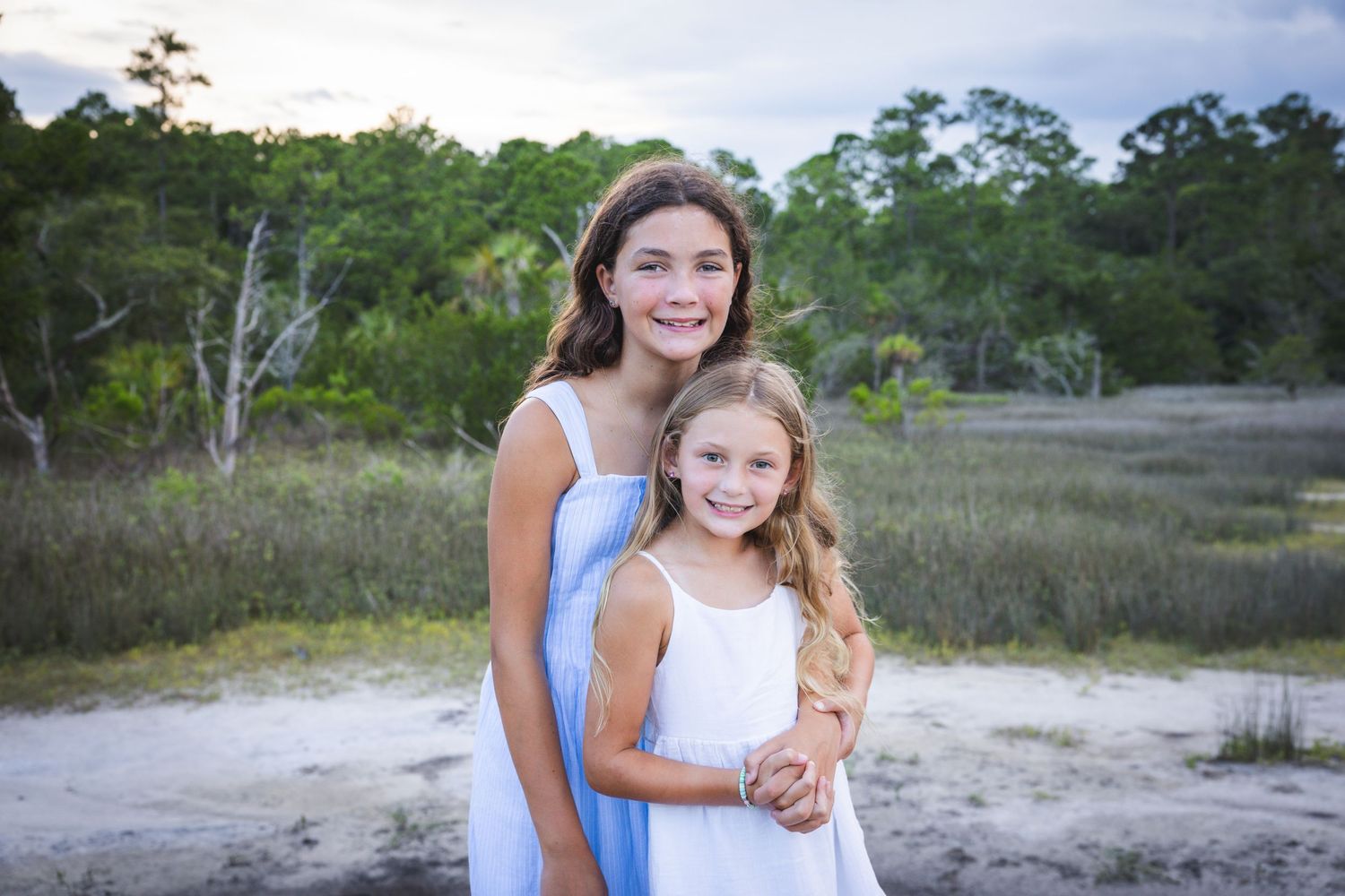 Two people in white and blue dresses embrace while standing in a natural coastal landscape at sunset.