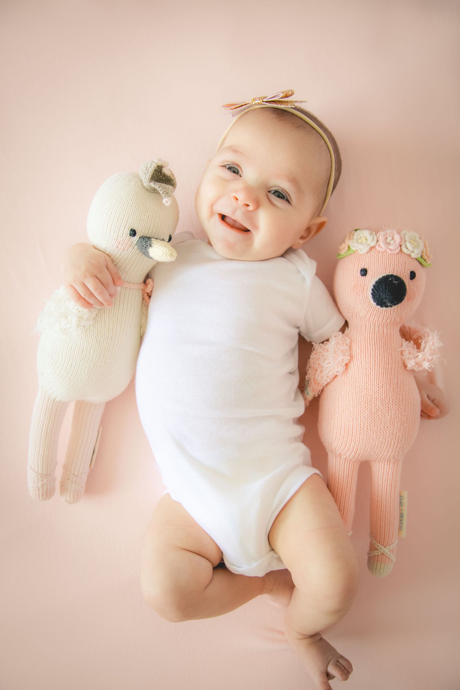 Smiling baby in white onesie lying between two knitted stuffed animals on pink background.