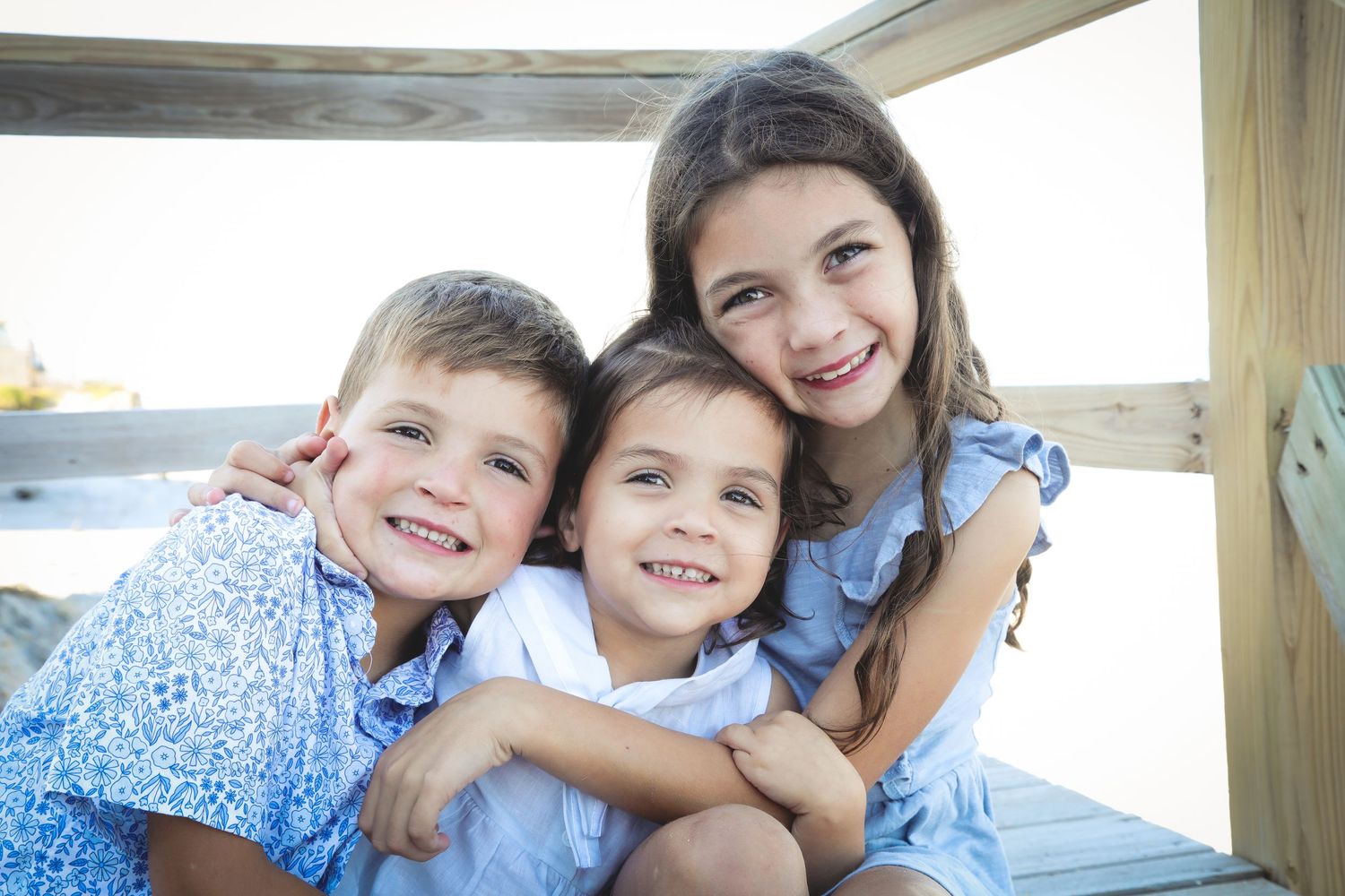Three children in blue clothing pose together on outdoor wooden deck.
