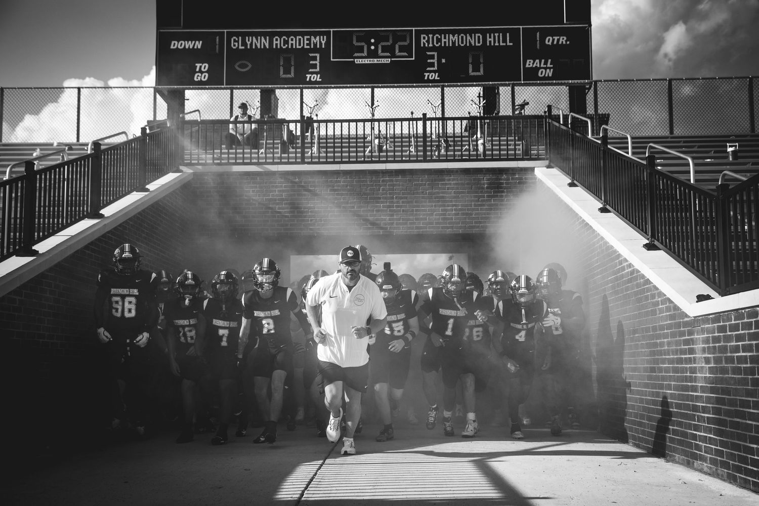 Black and white photo of a football stadium scoreboard with bleachers visible in background.