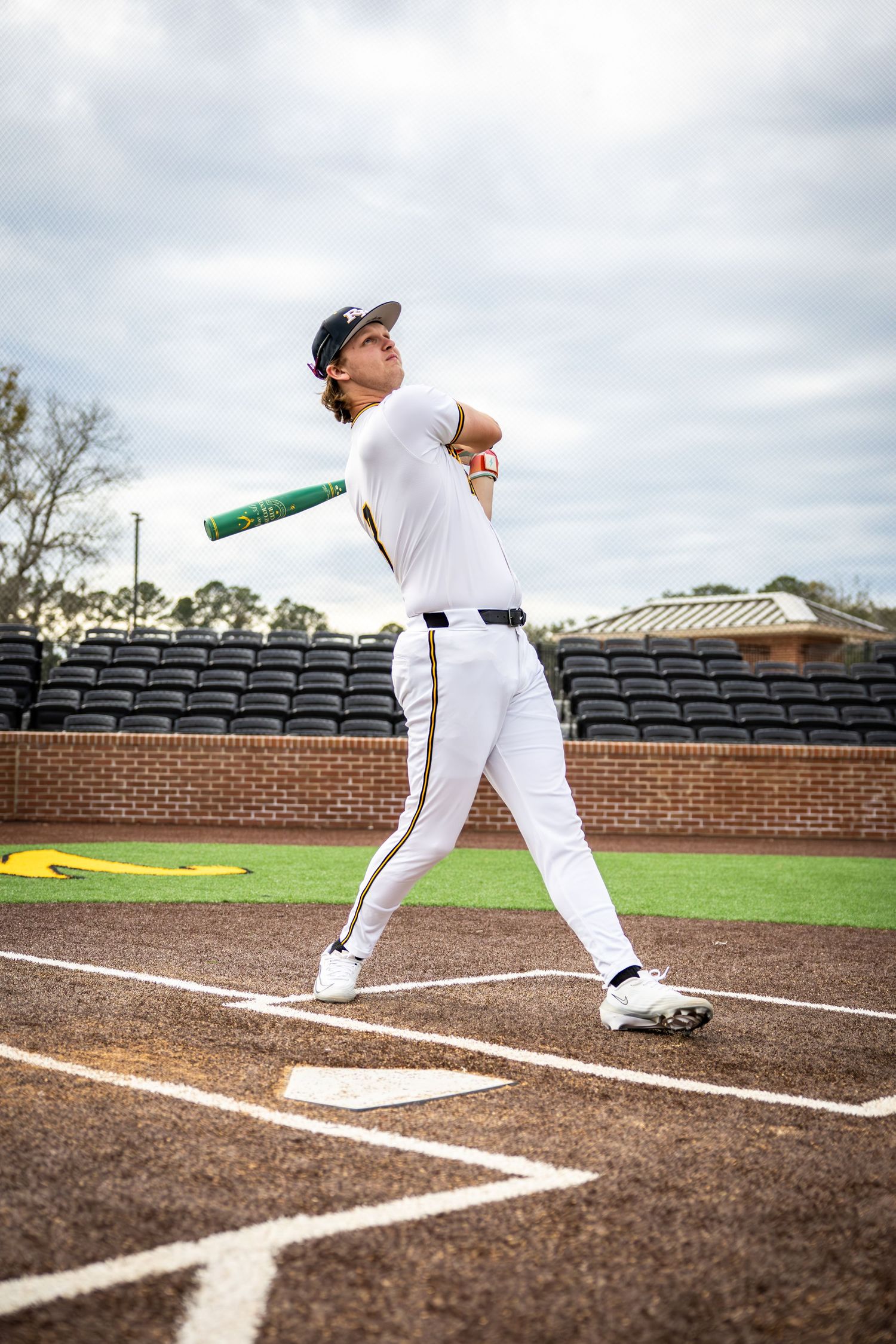 Baseball player in white uniform swings bat at home plate in an empty stadium on a cloudy day.