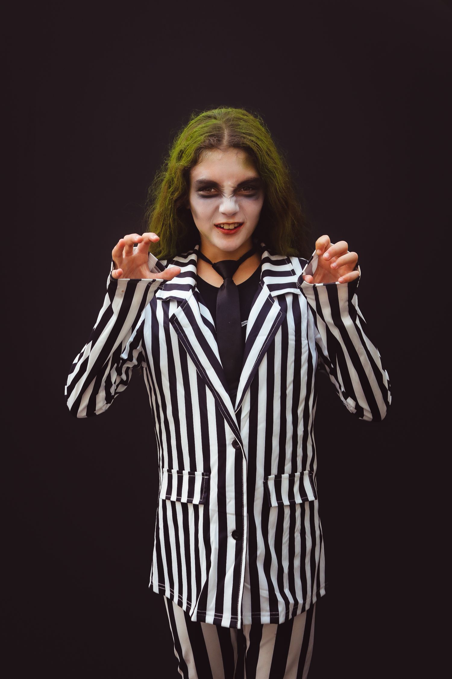 Person in black and white striped blazer with green hair in a spooky Halloween costume poses with raised hands.