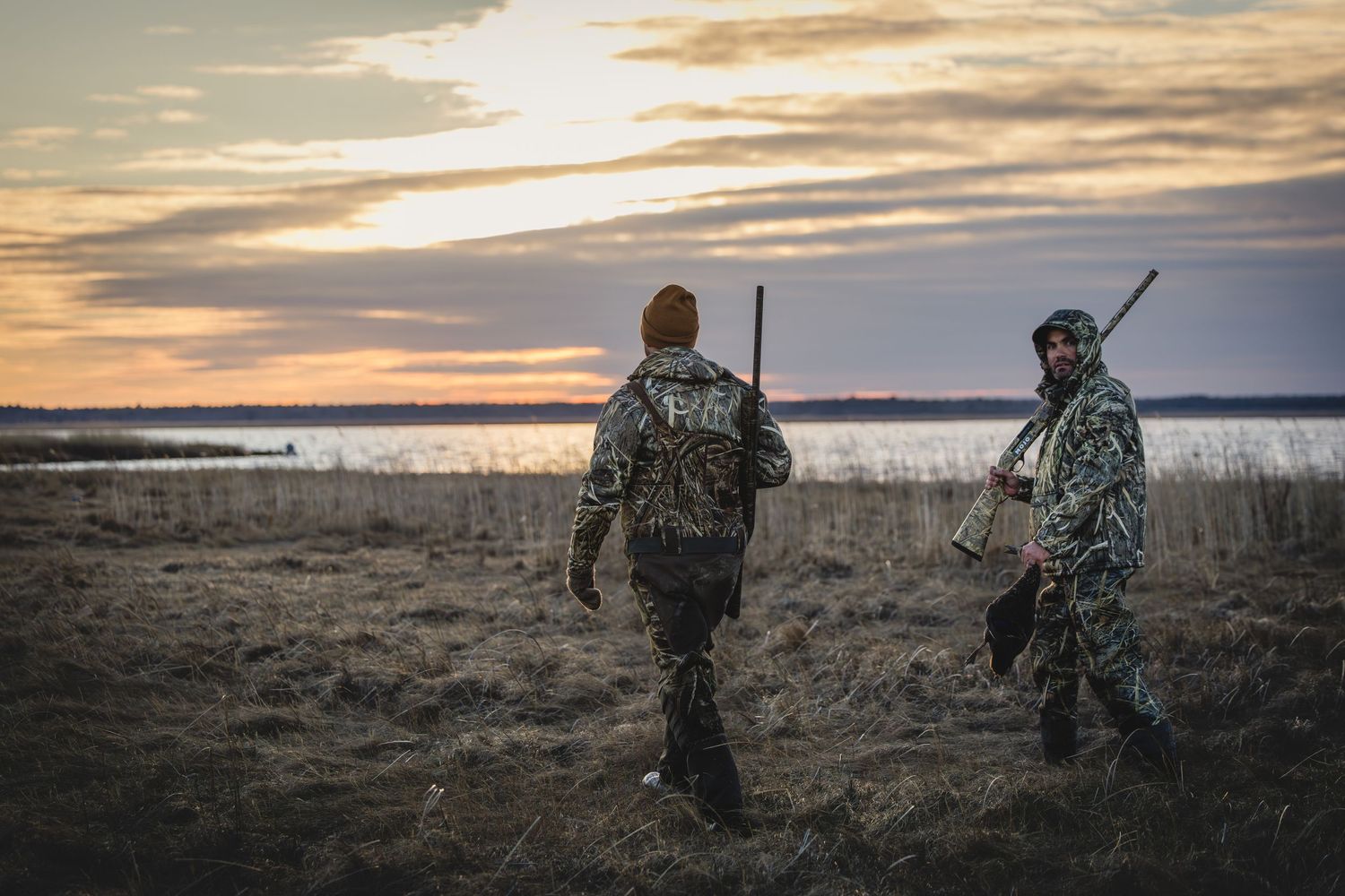 Hunters in camouflage gear walk through a field at sunset while waterfowl hunting near a lake.