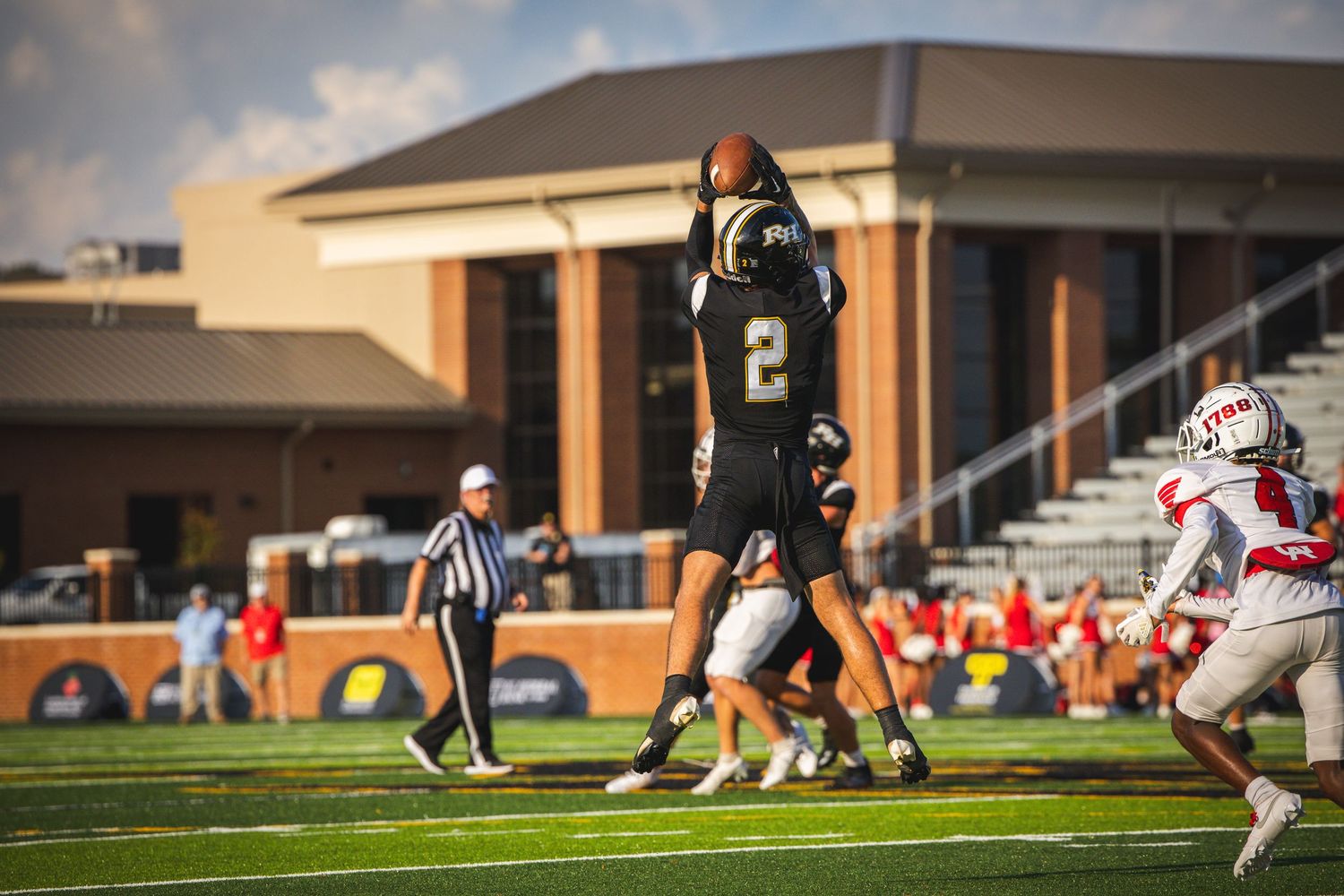 Football player #2 in black uniform leaps to catch ball during competitive game with referee and red team visible.