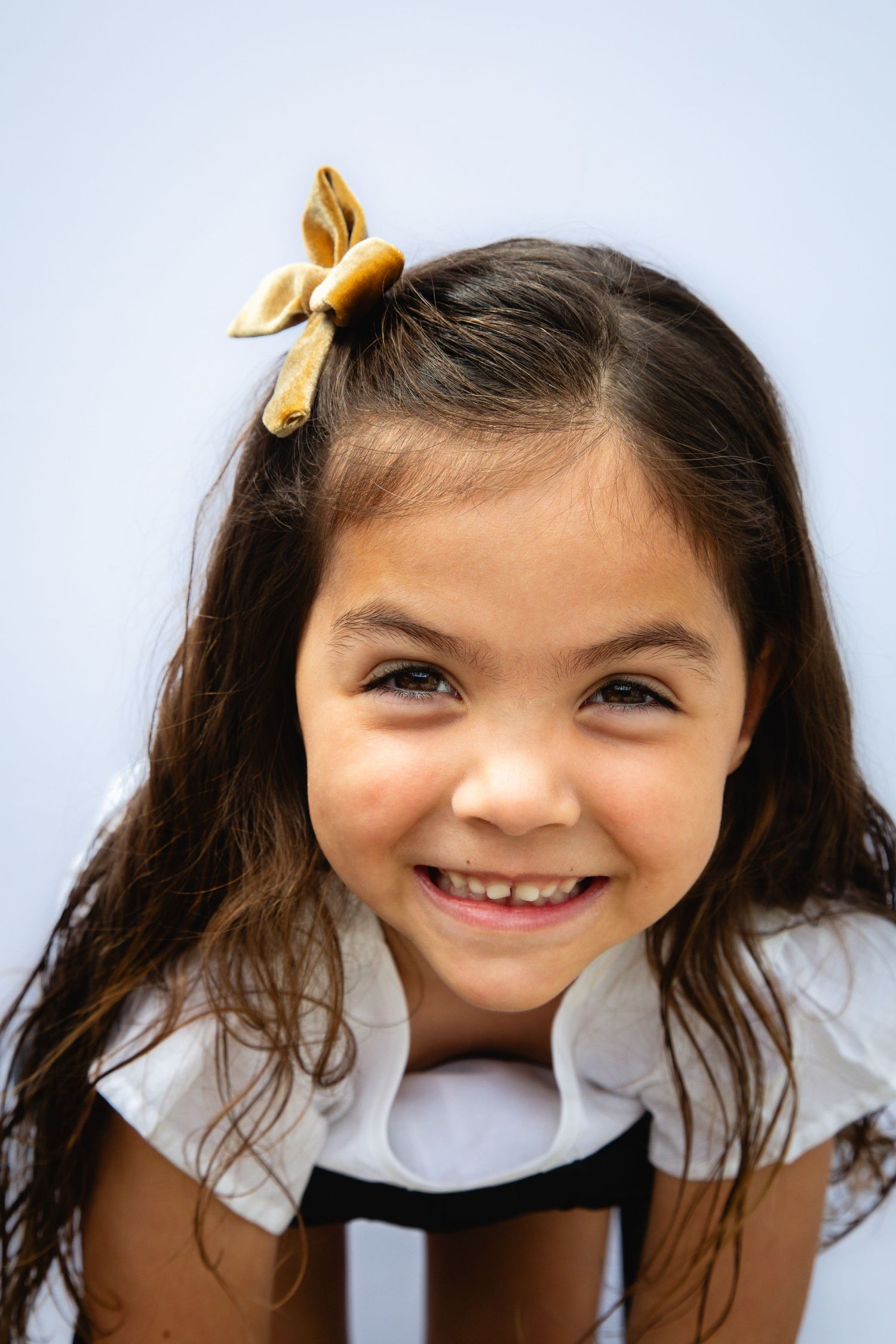 A young child with long dark hair wearing a mustard yellow bow and white top smiles brightly at the camera.