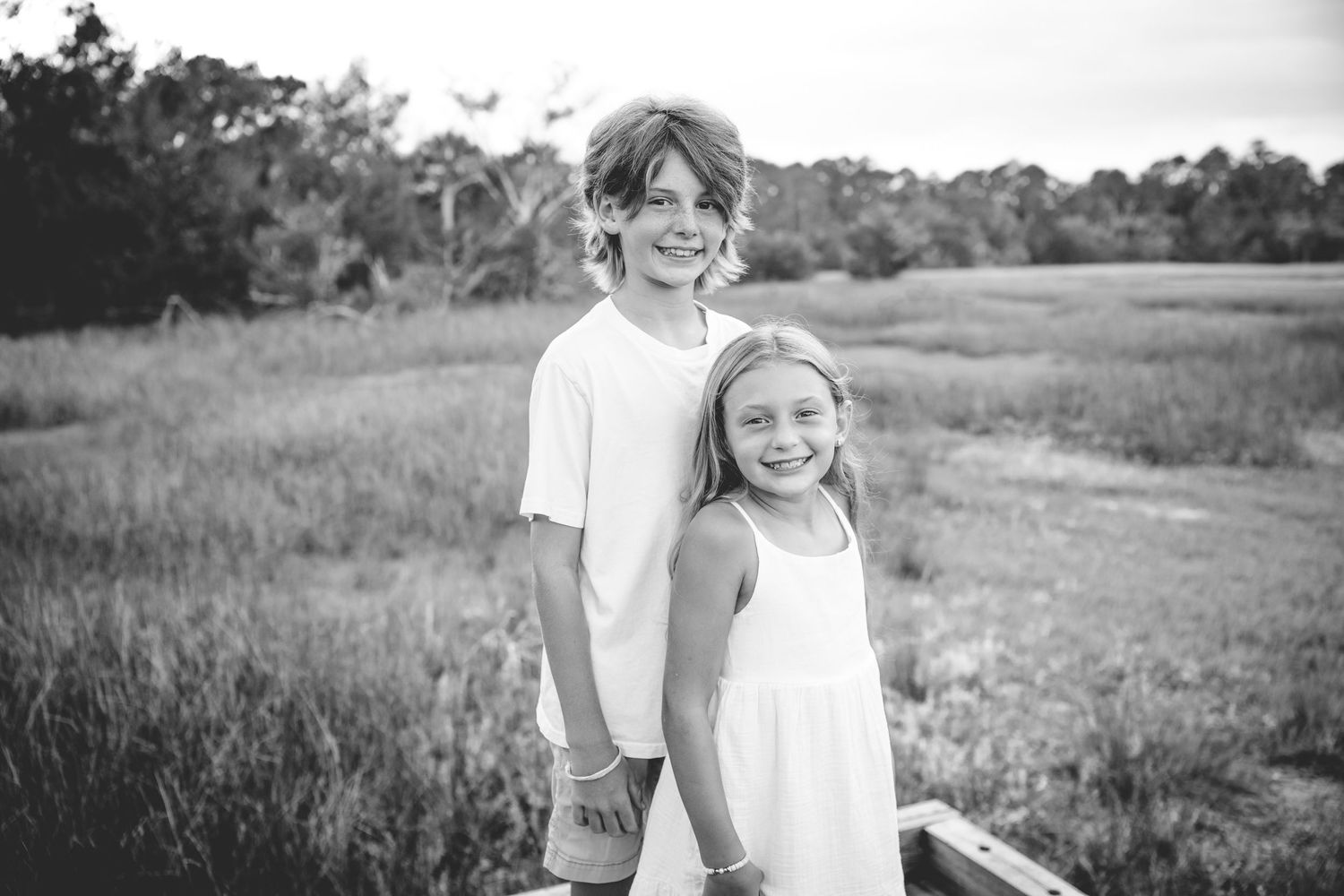 Two children in white outfits smile together on a boardwalk with marshland in the background.