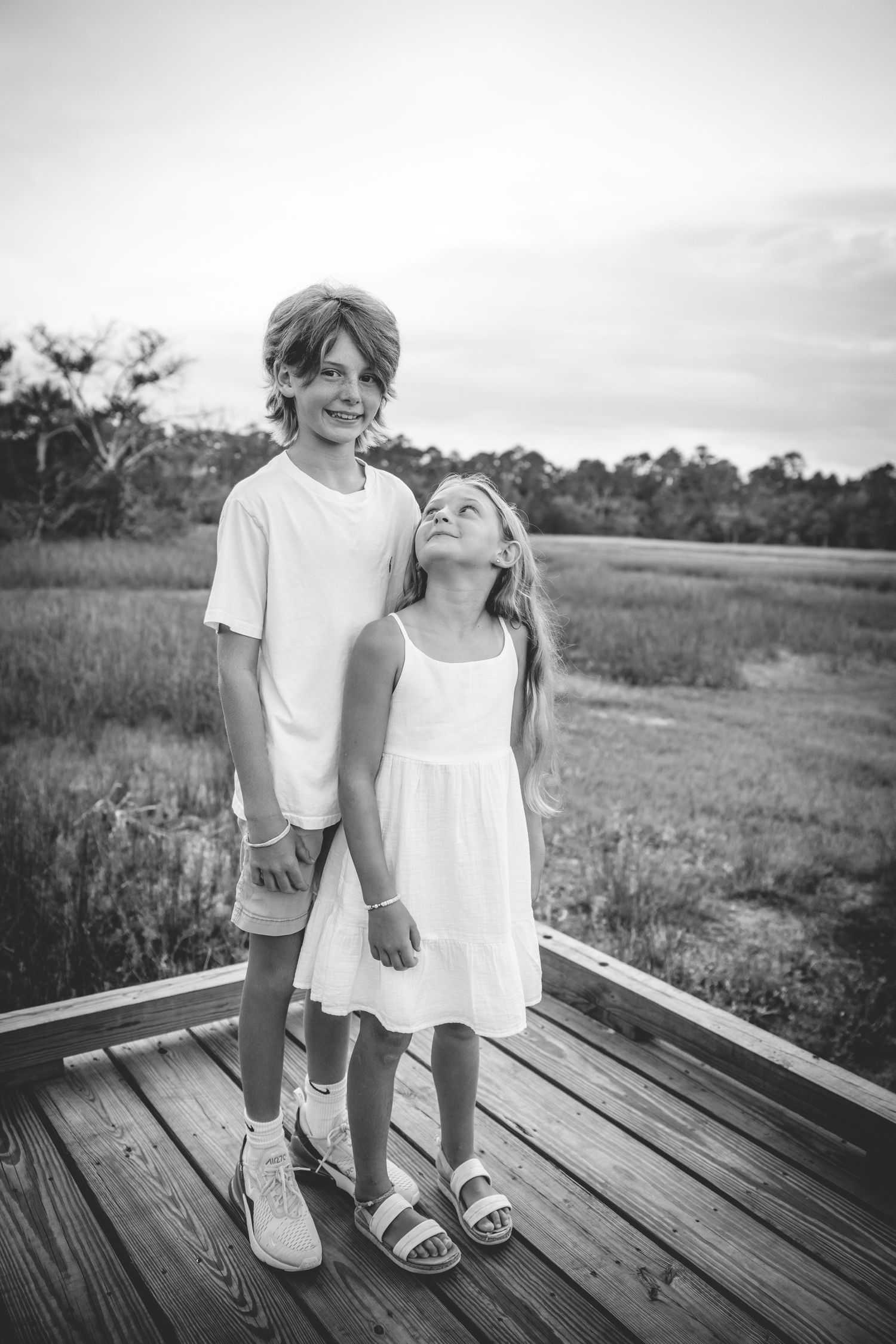 Siblings standing on wooden deck in a black and white photo with marsh and trees in background.