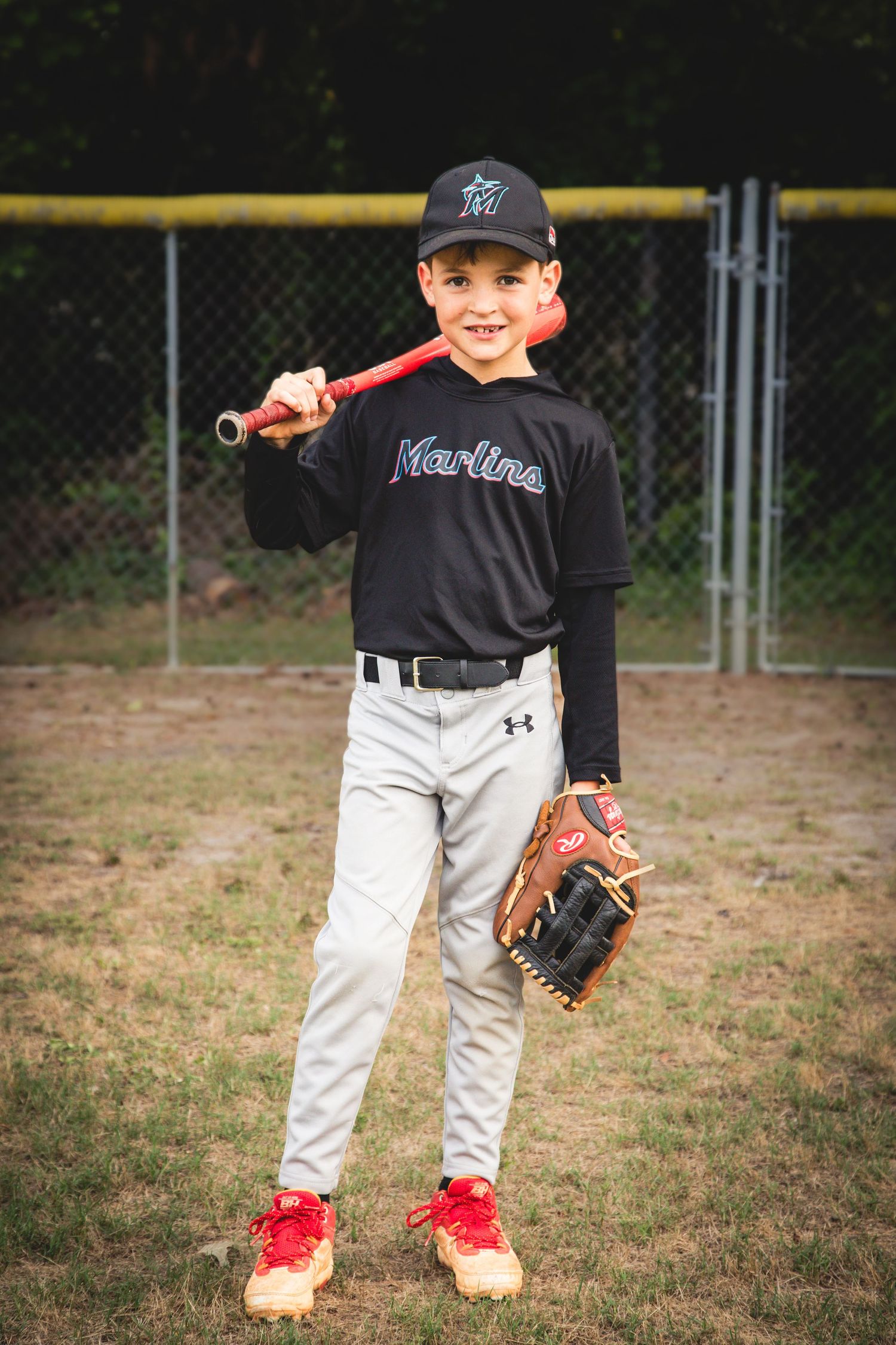 Youth baseball player in a black Marlins jersey and white pants poses with bat and glove on baseball field.
