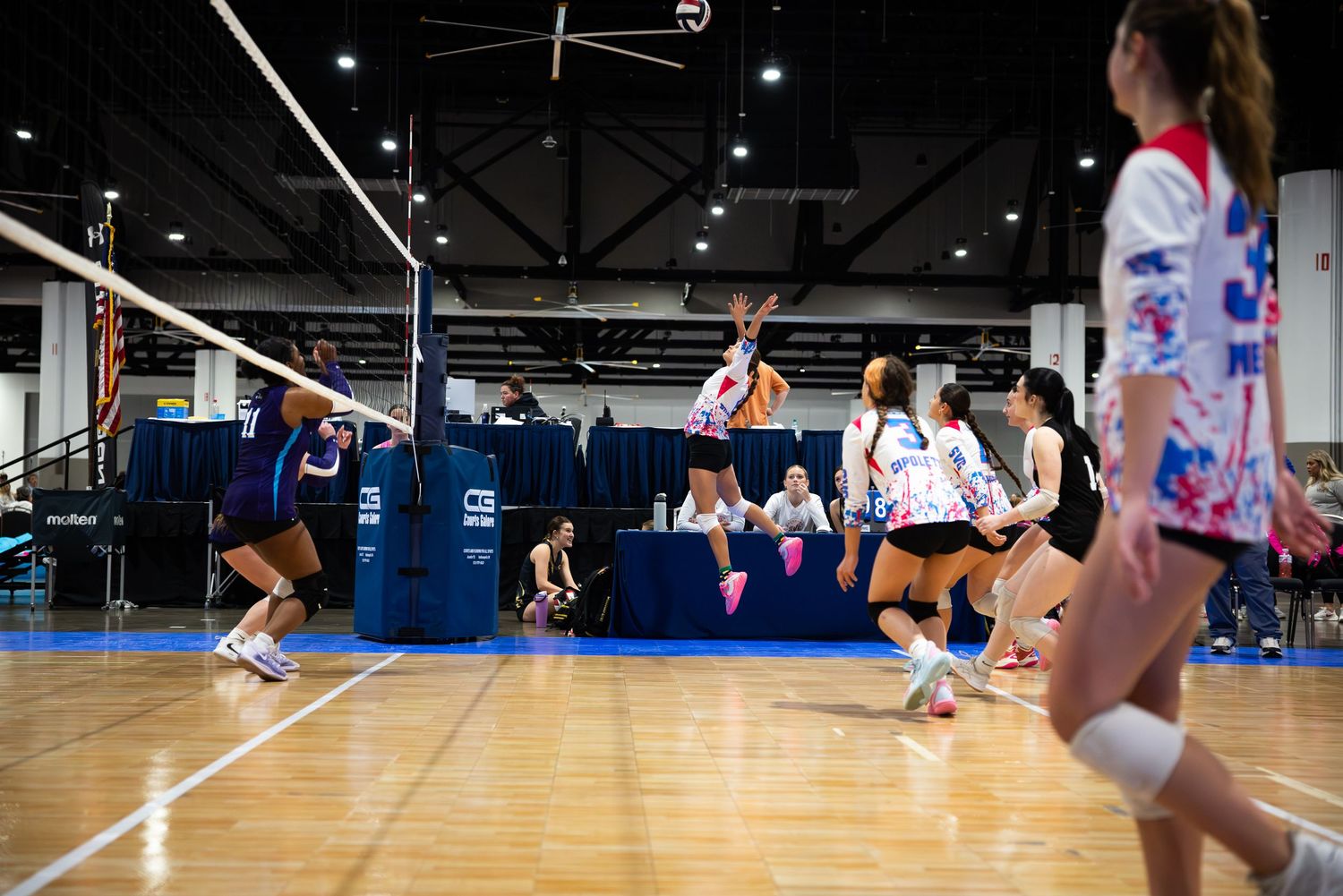 Volleyball players compete during an intense indoor tournament match with a player jumping to spike the ball.