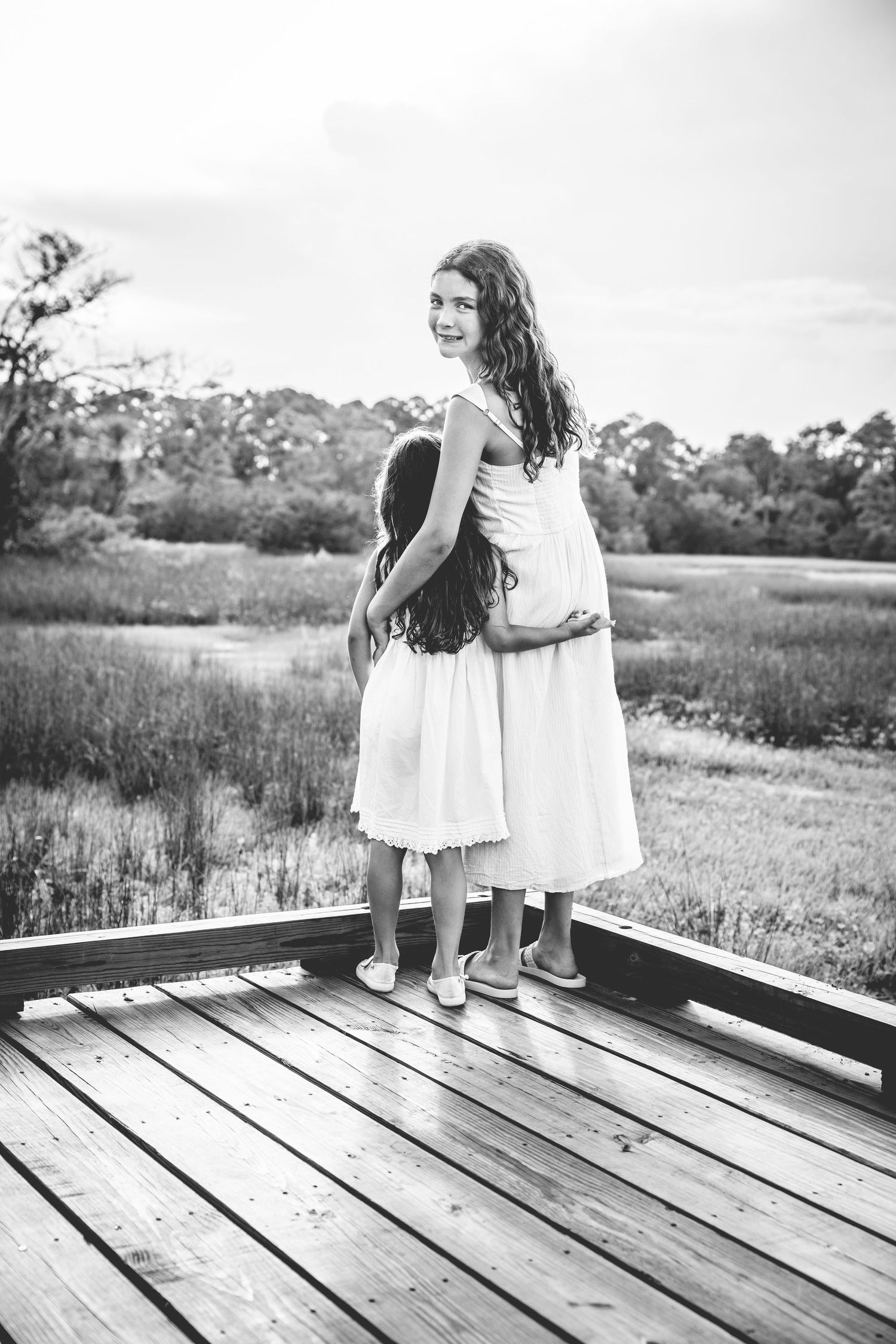 Black and white image of two children in white dresses holding hands and exploring a wooden boardwalk in nature.