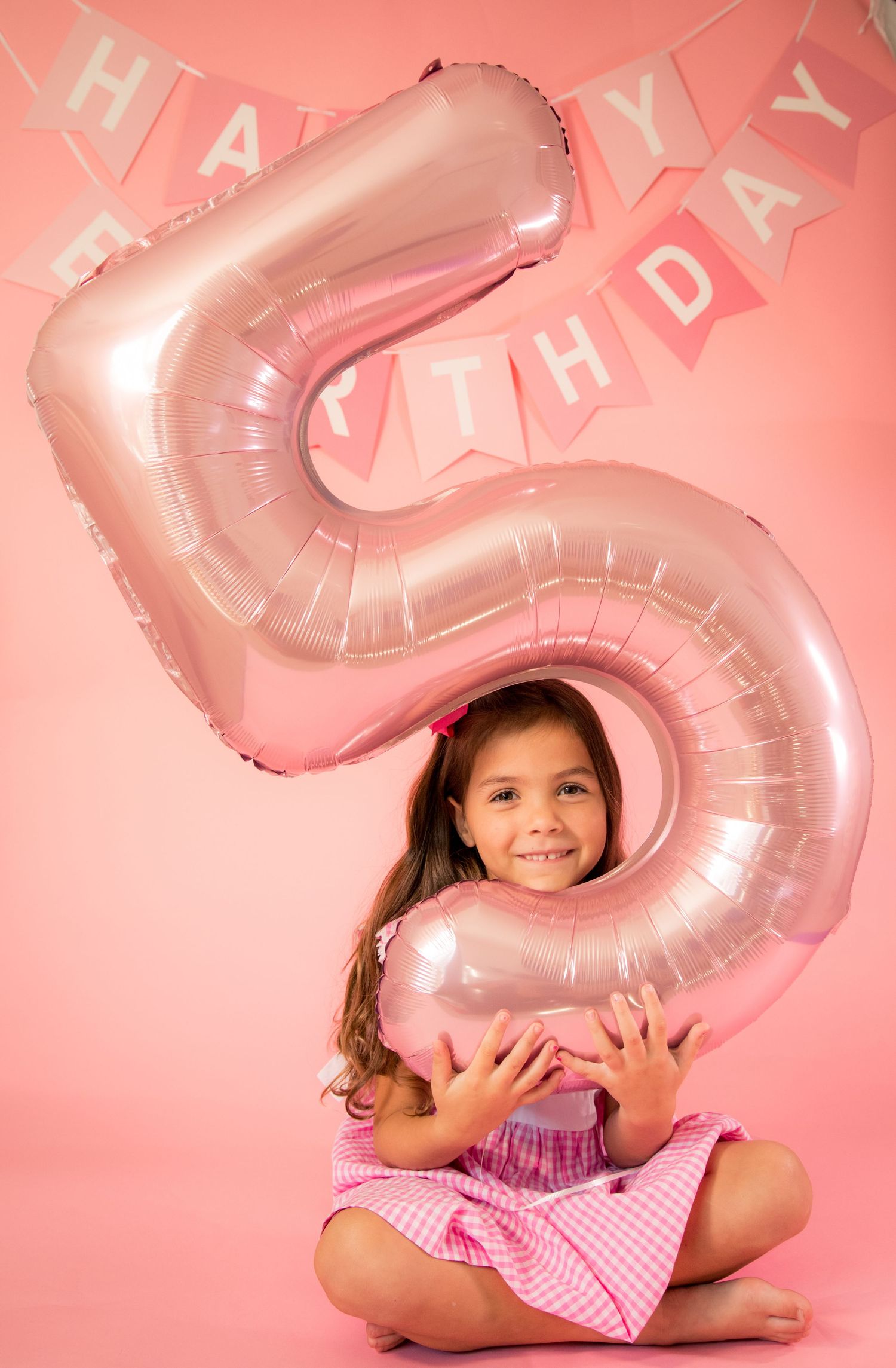 Child in pink and white gingham dress poses with large silver balloon numbers against pink background.