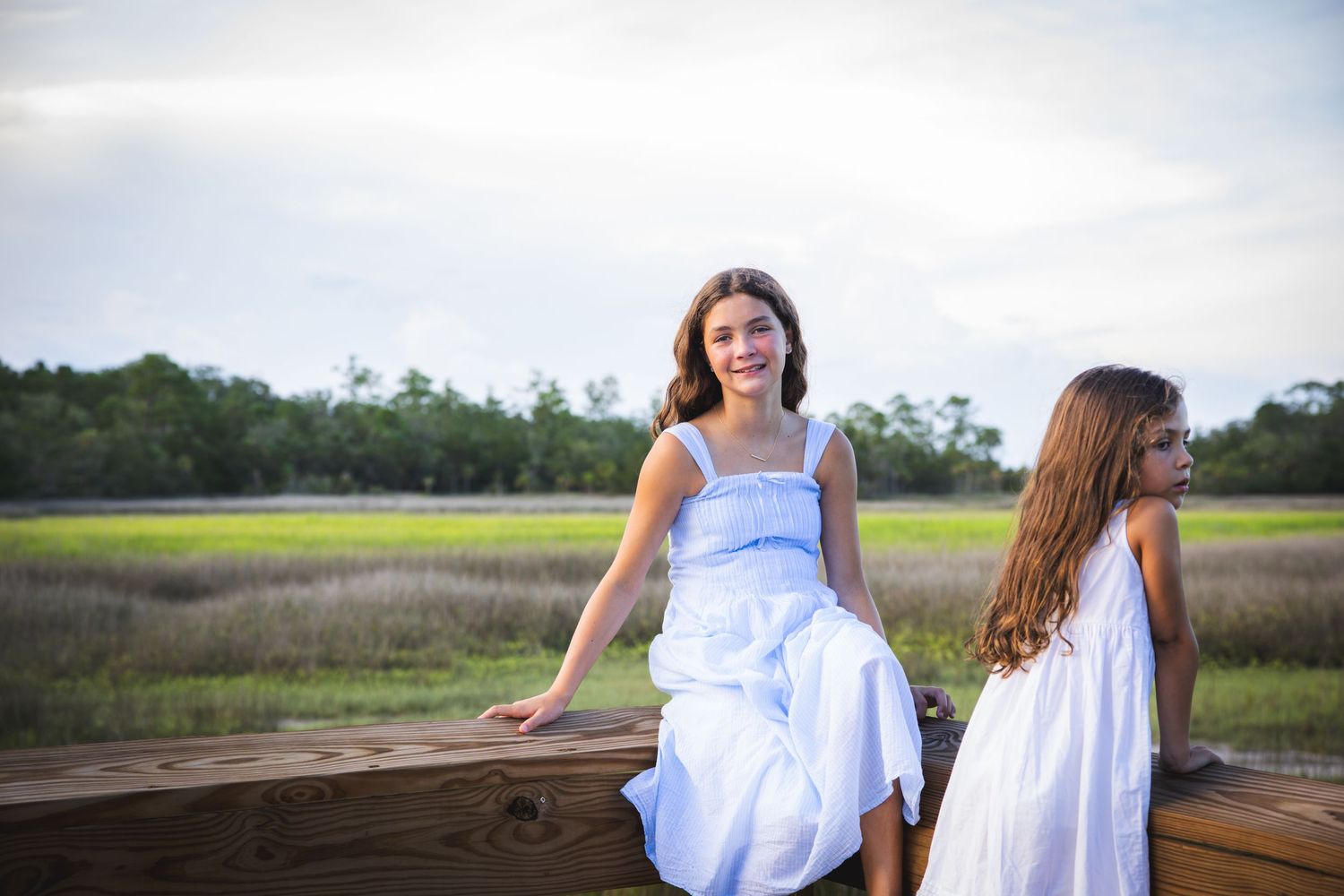 Two people in white dresses sit on a wooden deck overlooking a marshy landscape at sunset.