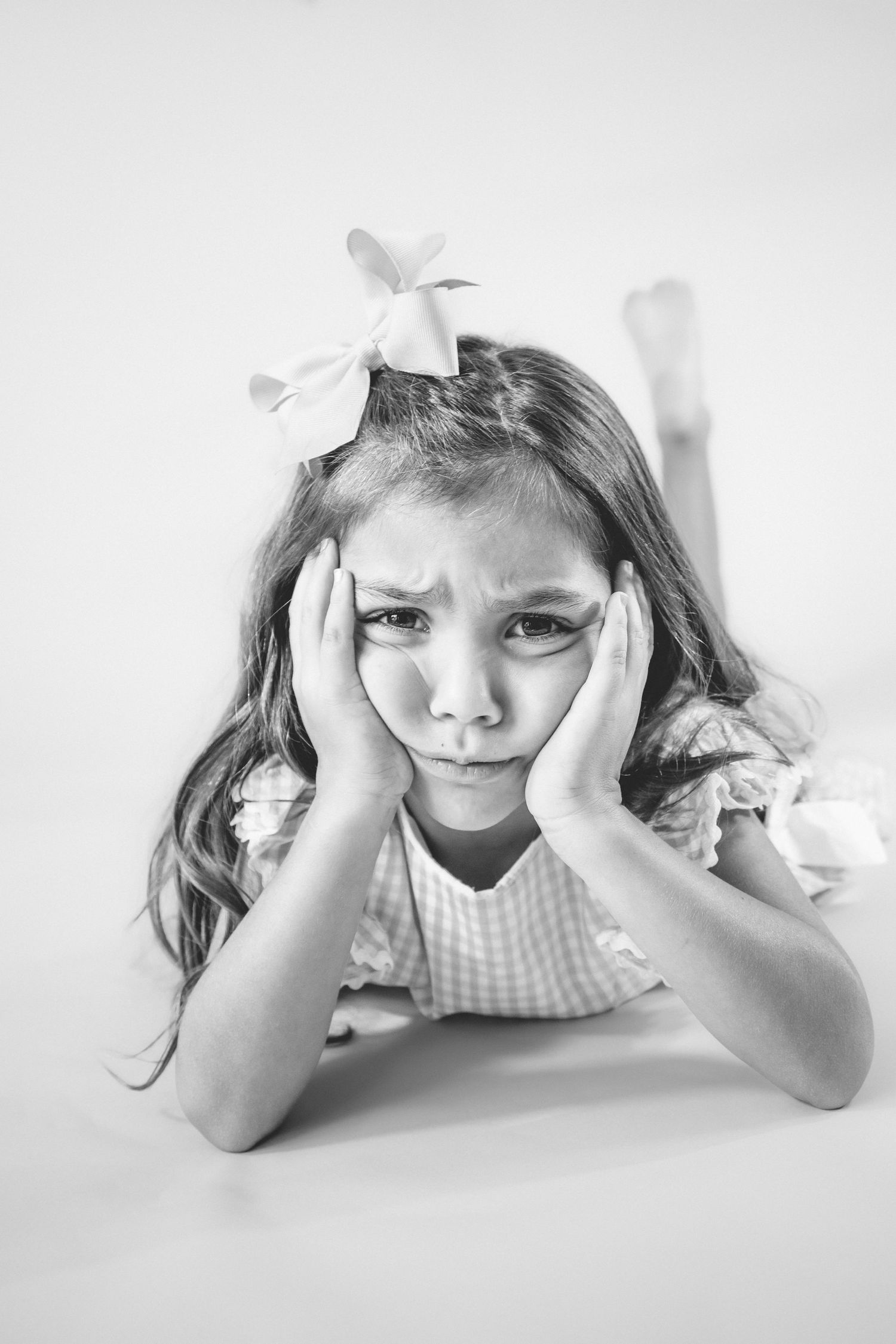 A black and white portrait of a young girl in a gingham dress lying down with hands on face and bow in her hair.