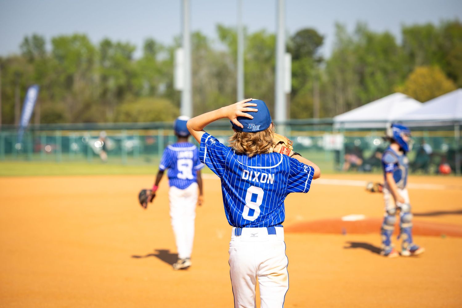 Youth baseball player #8 Dixon walks on a sunny dirt field in a blue uniform, hand on head, during a little league game.