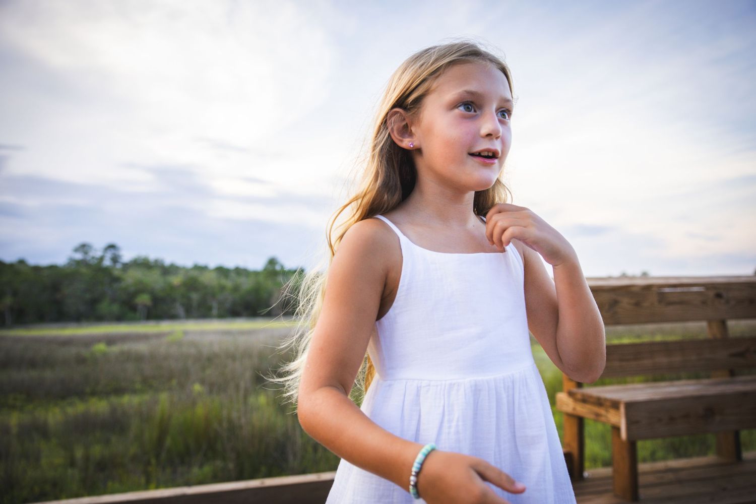 Person in a white dress stands on a wooden deck overlooking a coastal marsh at dusk.