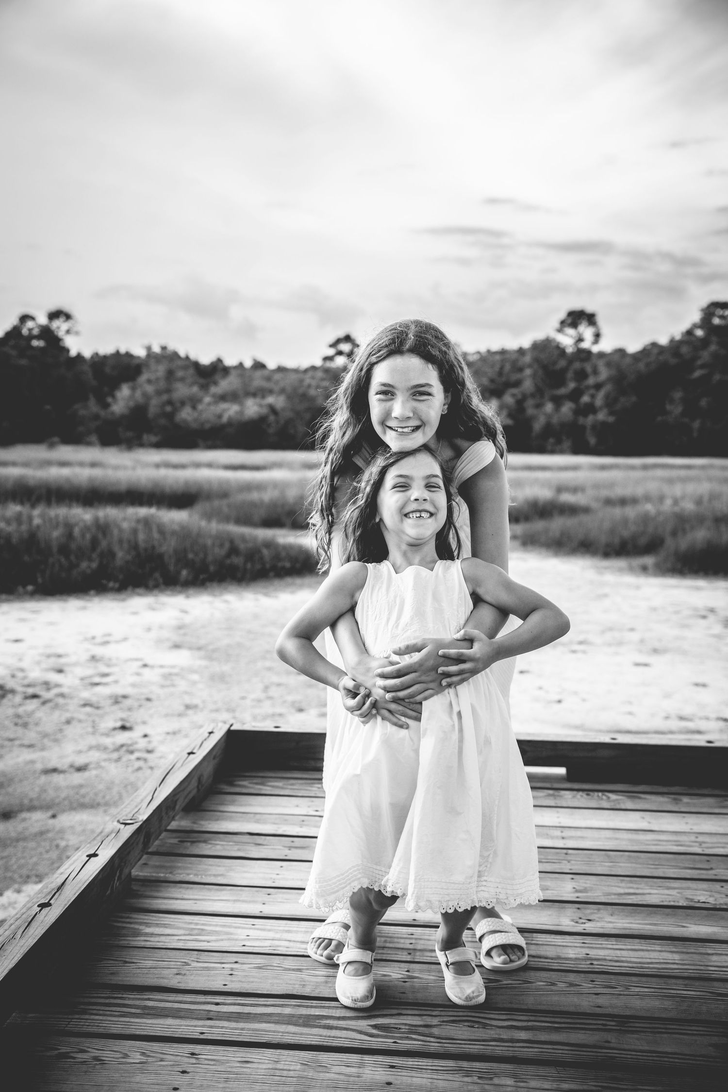 Two children embracing and smiling on a wooden dock with nature landscape behind them.