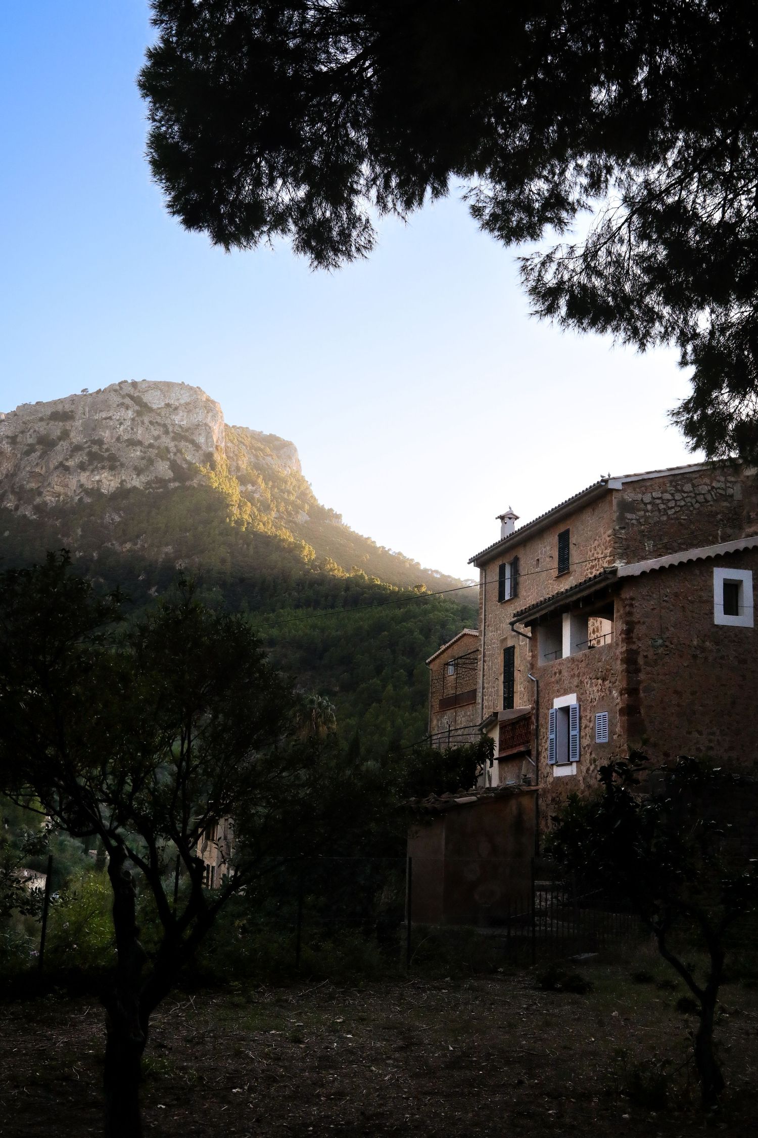 Mediterranean stone house nestled against mountain backdrop with blue sky and framing trees in Mallorca, Spain.