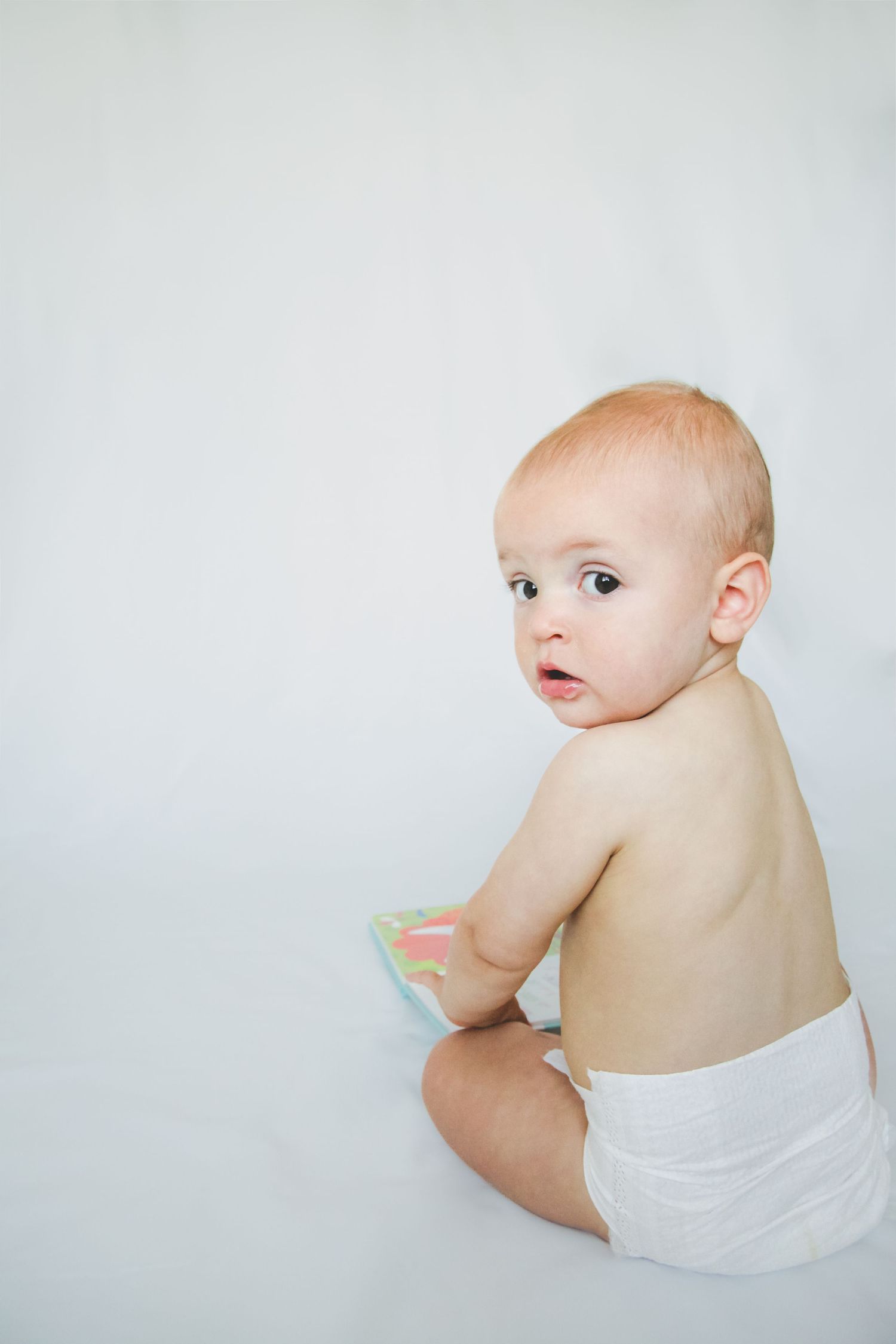 An infant wearing a white diaper sits on a light background while looking over their shoulder.