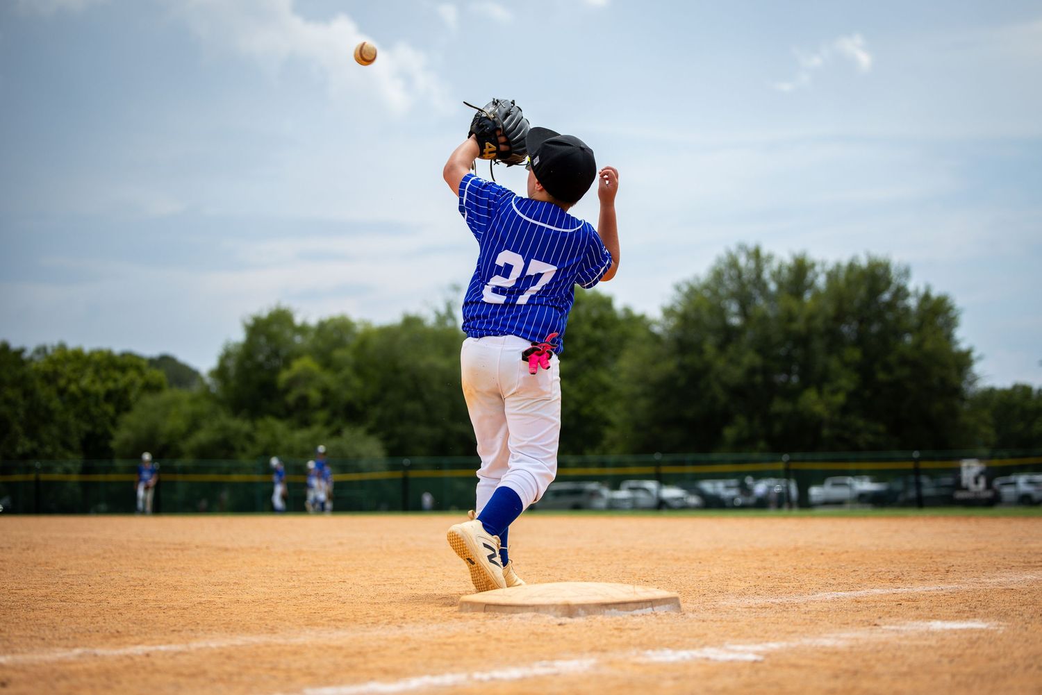 Youth baseball player #27 in blue uniform catching a fly ball at first base on a sunny day.