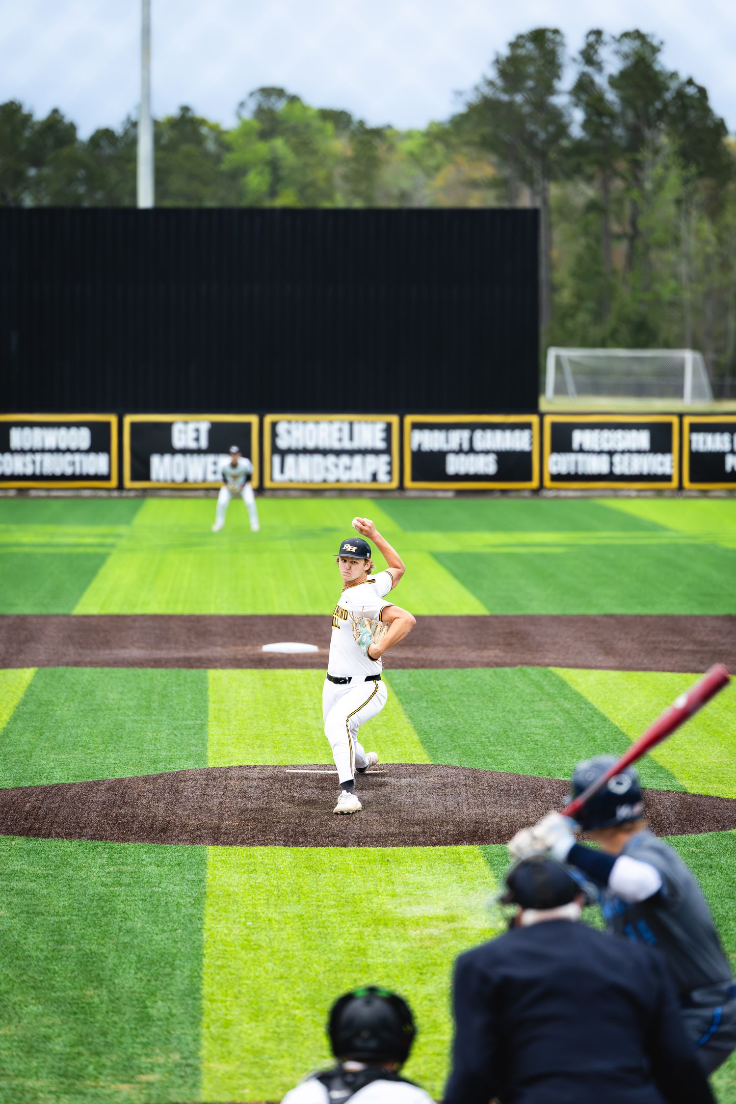 Baseball pitcher winds up on the mound during a game, with batter and catcher in foreground and green outfield behind.