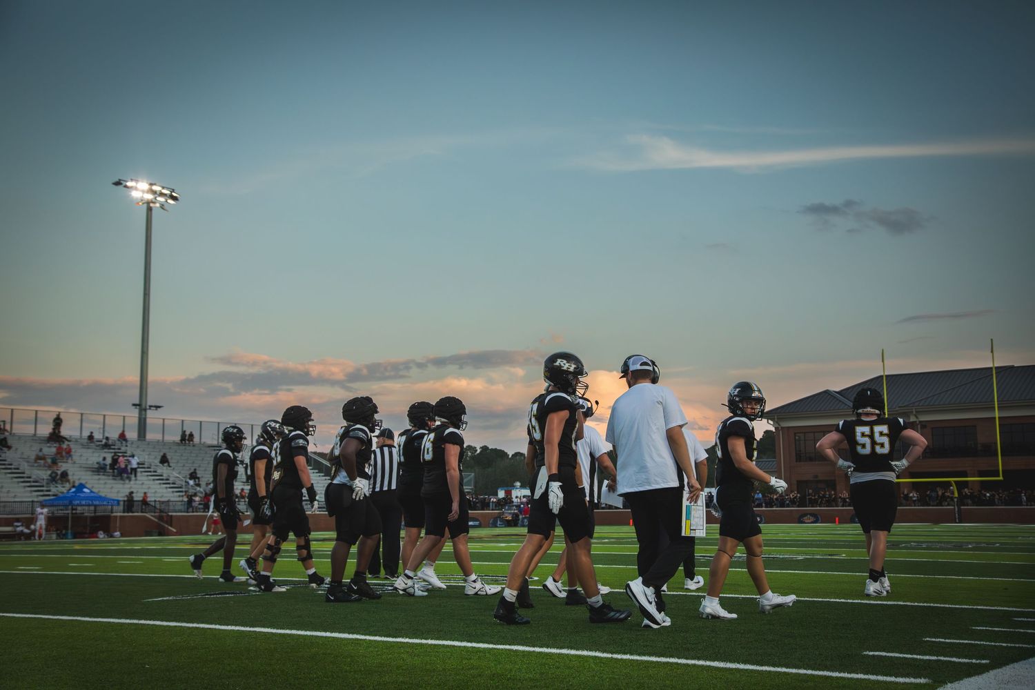 Football players line up during sunset on a turf field for preseason practice drills.