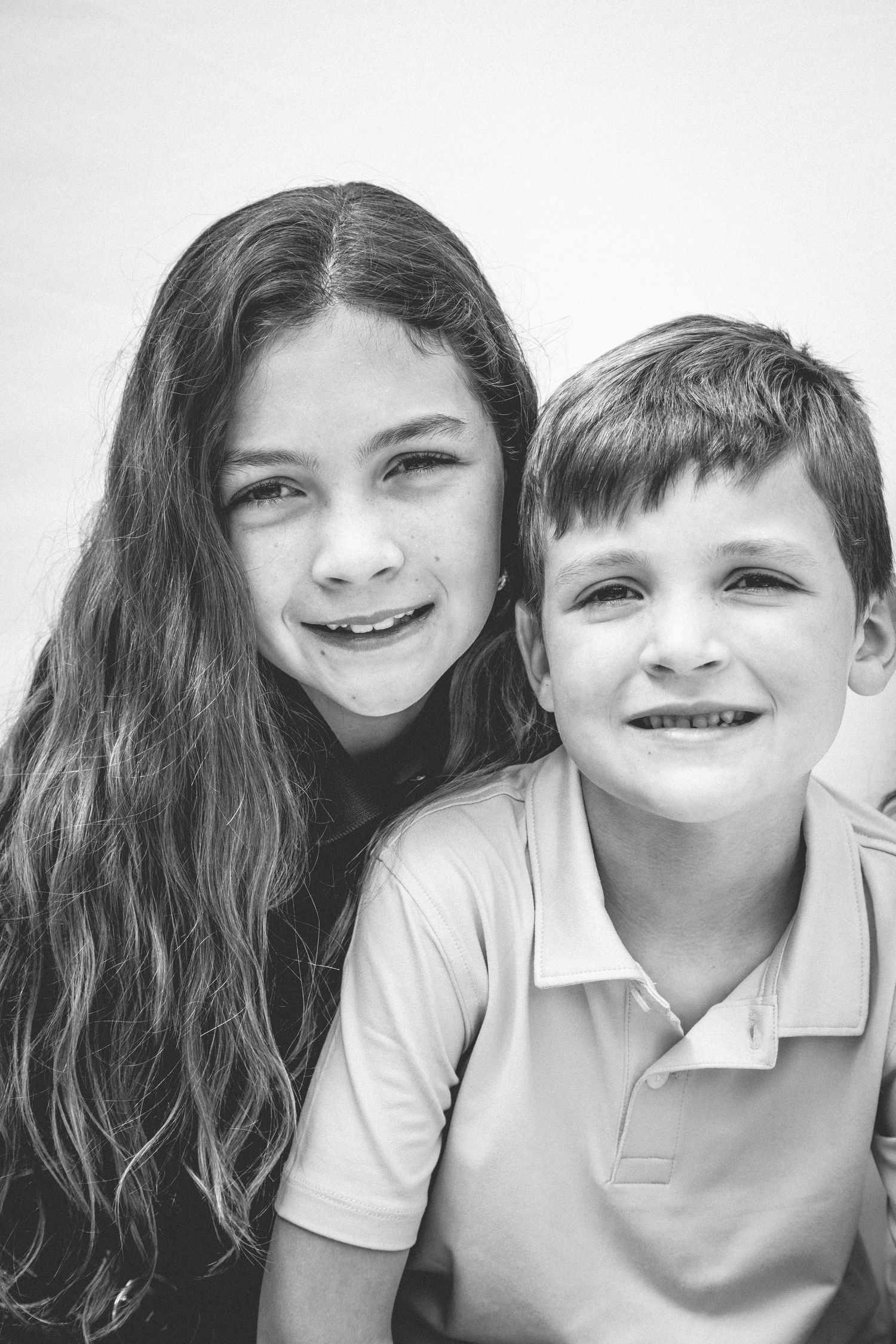 Two happy children in white polo shirts smile together in a black and white photo series.