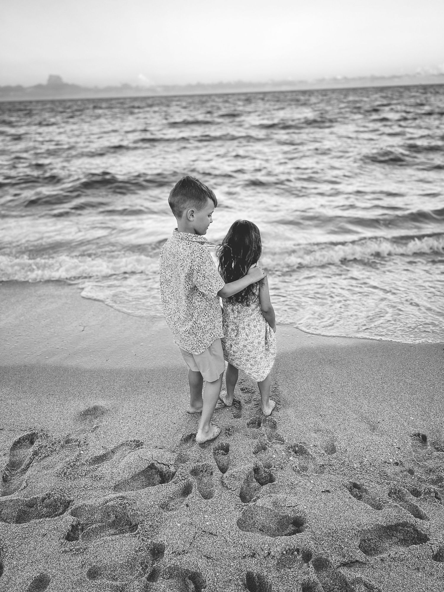 Two small children stand on a sandy beach embracing while looking out at the ocean waves and distant shore in black and white.