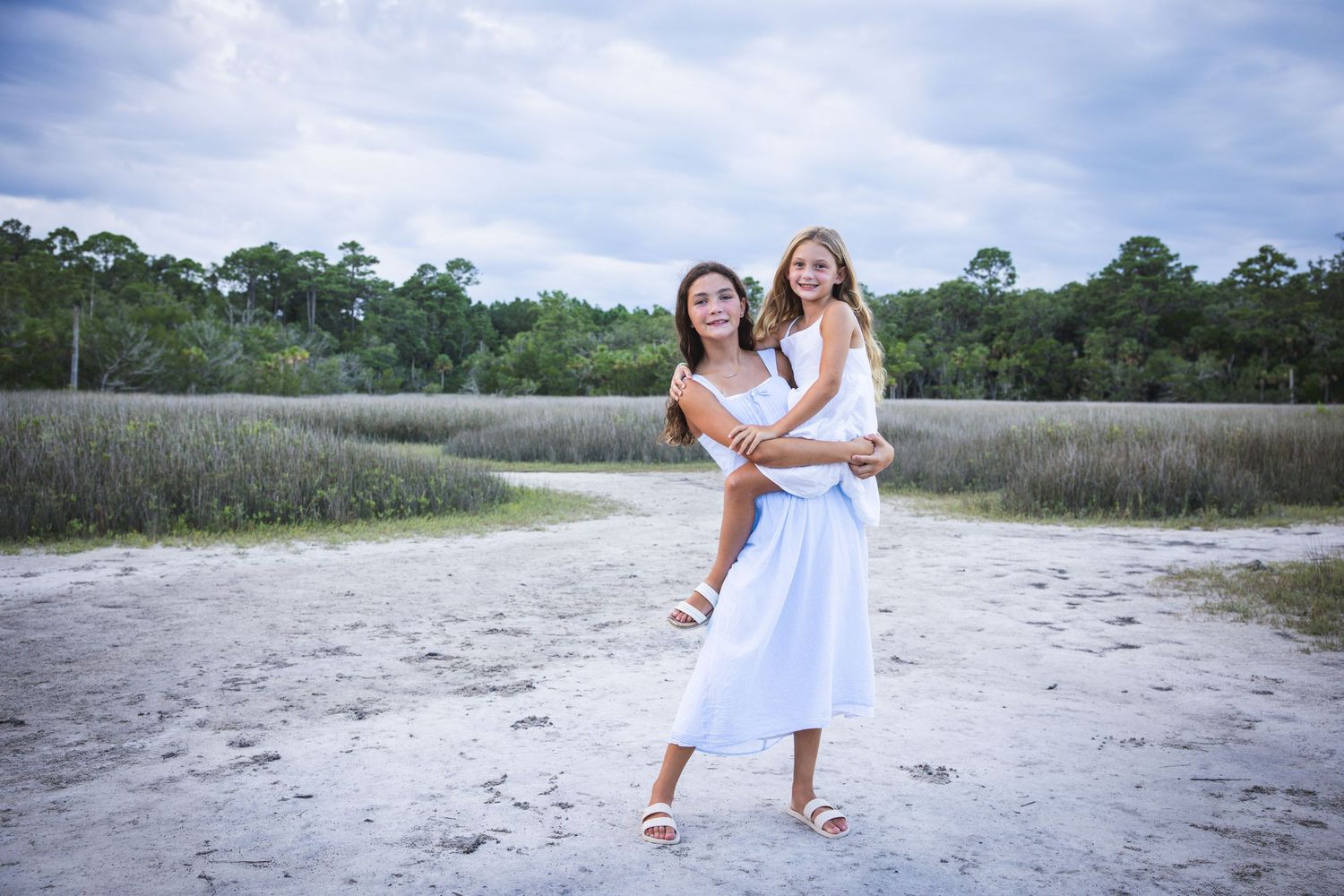 Two people in white dresses embrace while standing on sandy beach with marsh and trees in background.