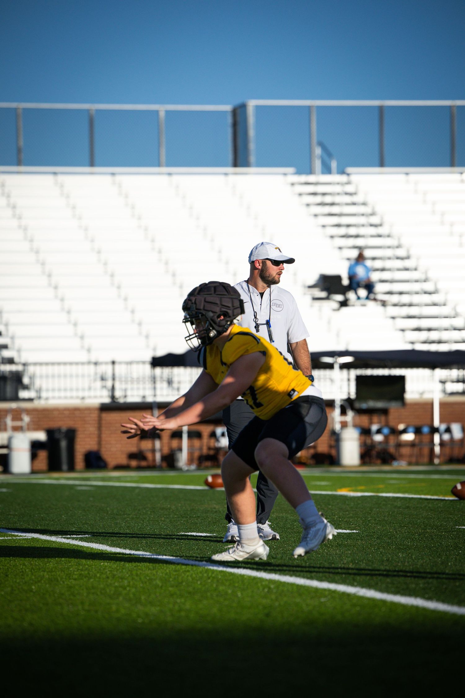 Football player in yellow jersey practices drills on field while coach observes during outdoor training session.