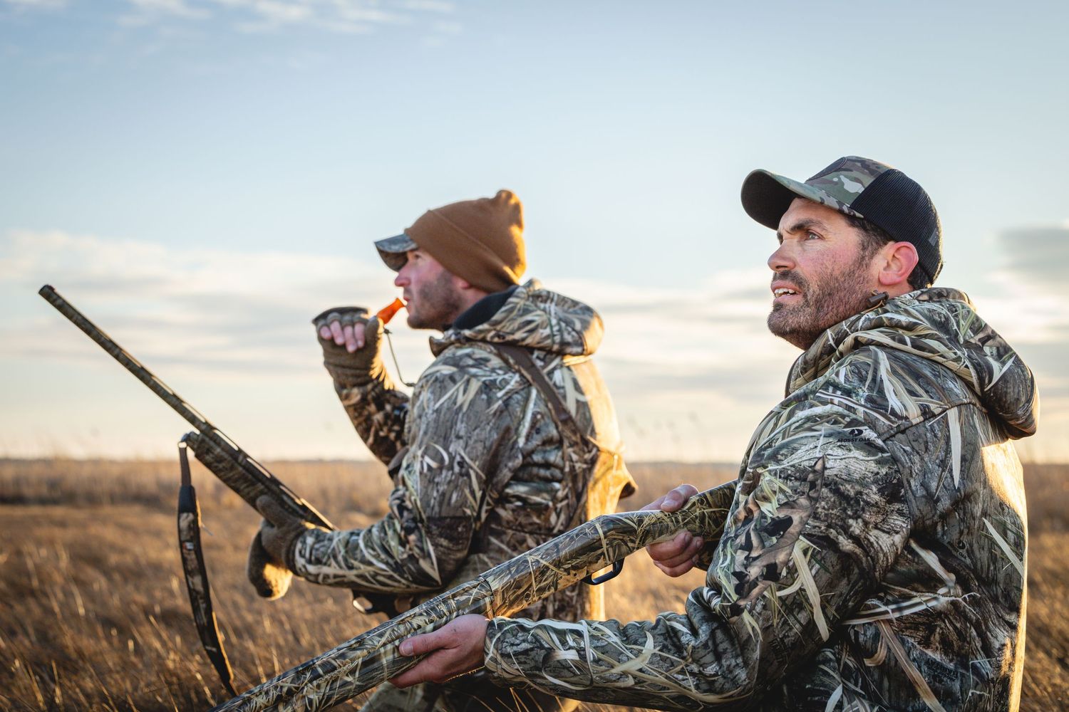 Hunters in camouflage gear scanning the horizon with binoculars at sunset in an open field.