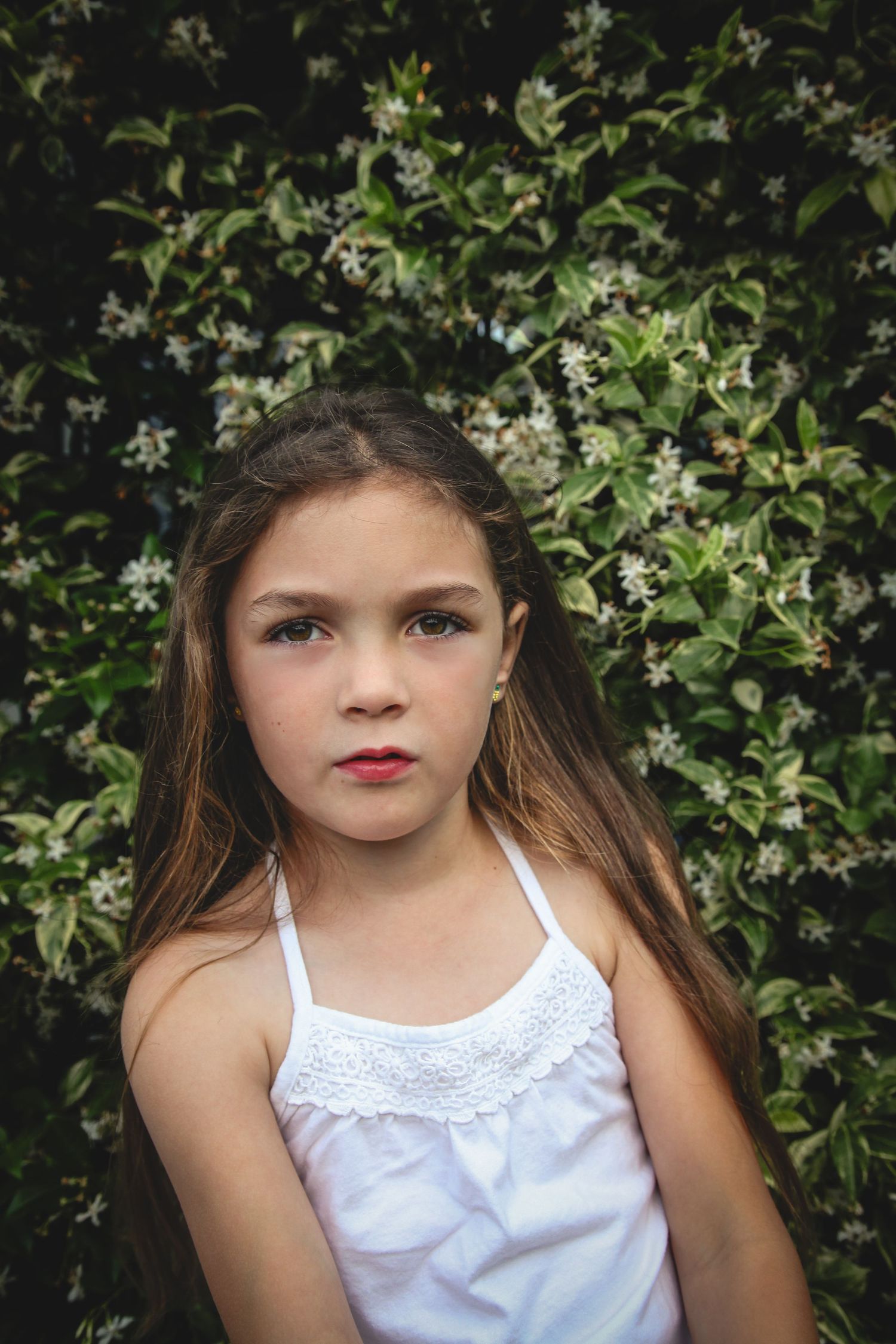 Young performer in a white lace tank top poses against a flowering bush backdrop in soft lighting.