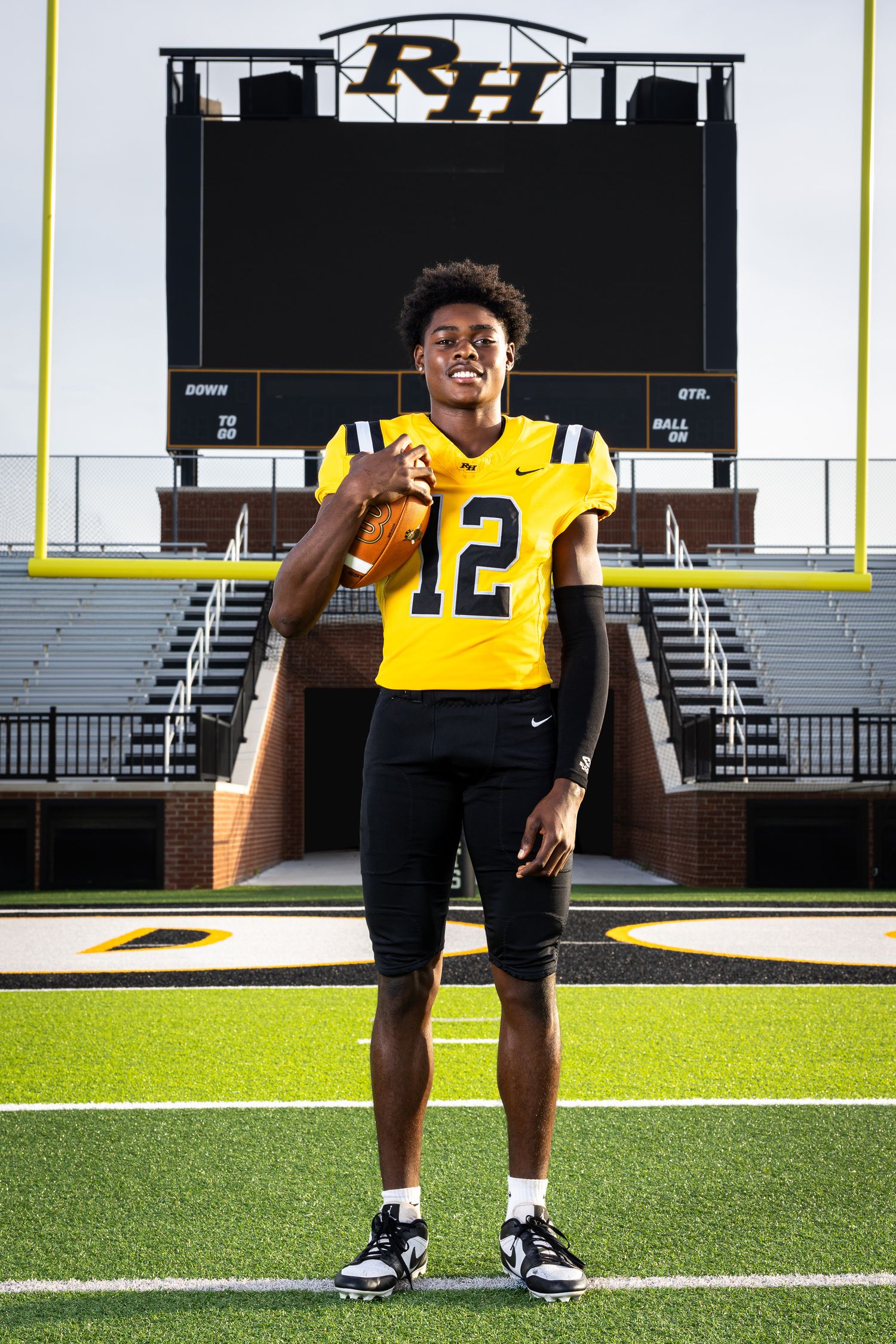 High school football player #12 poses in gold and black uniform on field with RH scoreboard backdrop.