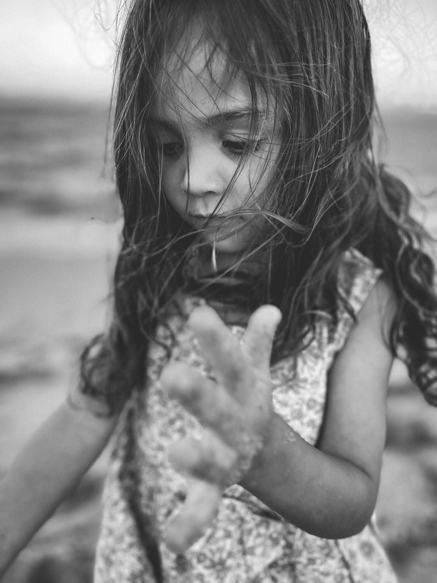 Black and white artistic portrait of a small child with windswept hair wearing a floral dress outdoors.