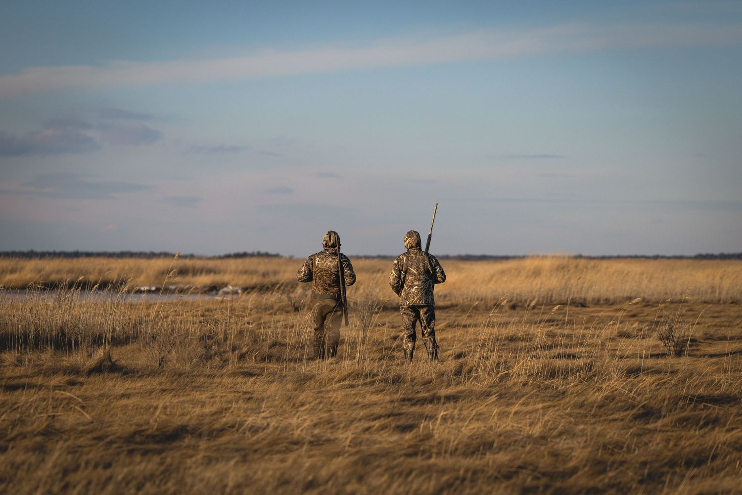 Hunters walking through tall golden prairie grass at sunset with rifles and hunting gear in a vast open landscape.