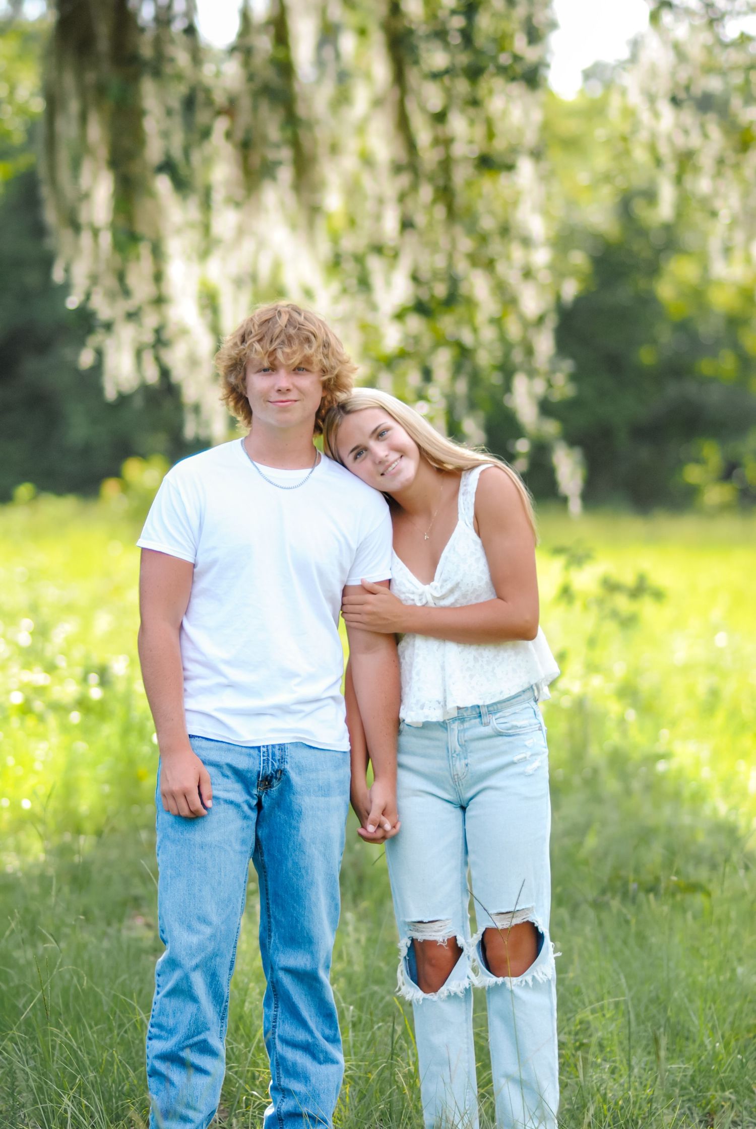 A couple in casual denim and white outfits poses together in a sunny meadow.